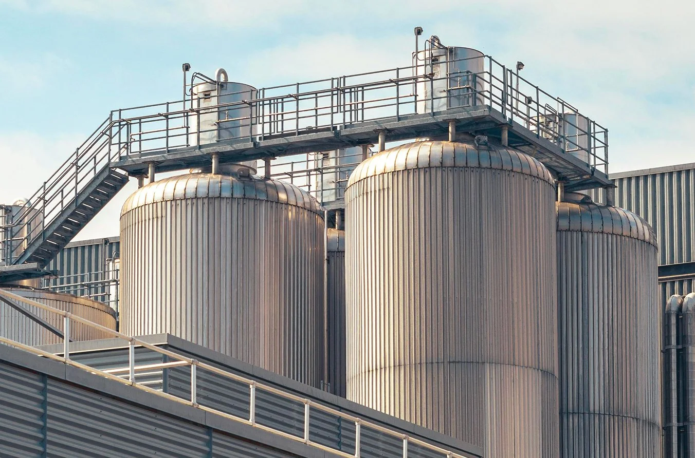 Industrial facility with large metallic storage tanks, piping, and railings on top, under a blue sky.