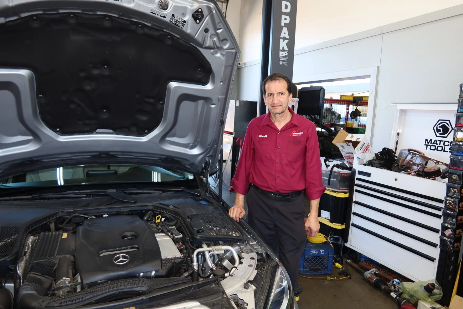 A mechanic in a red work shirt stands next to an open Mercedes-Benz engine hood inside an automotive repair shop.