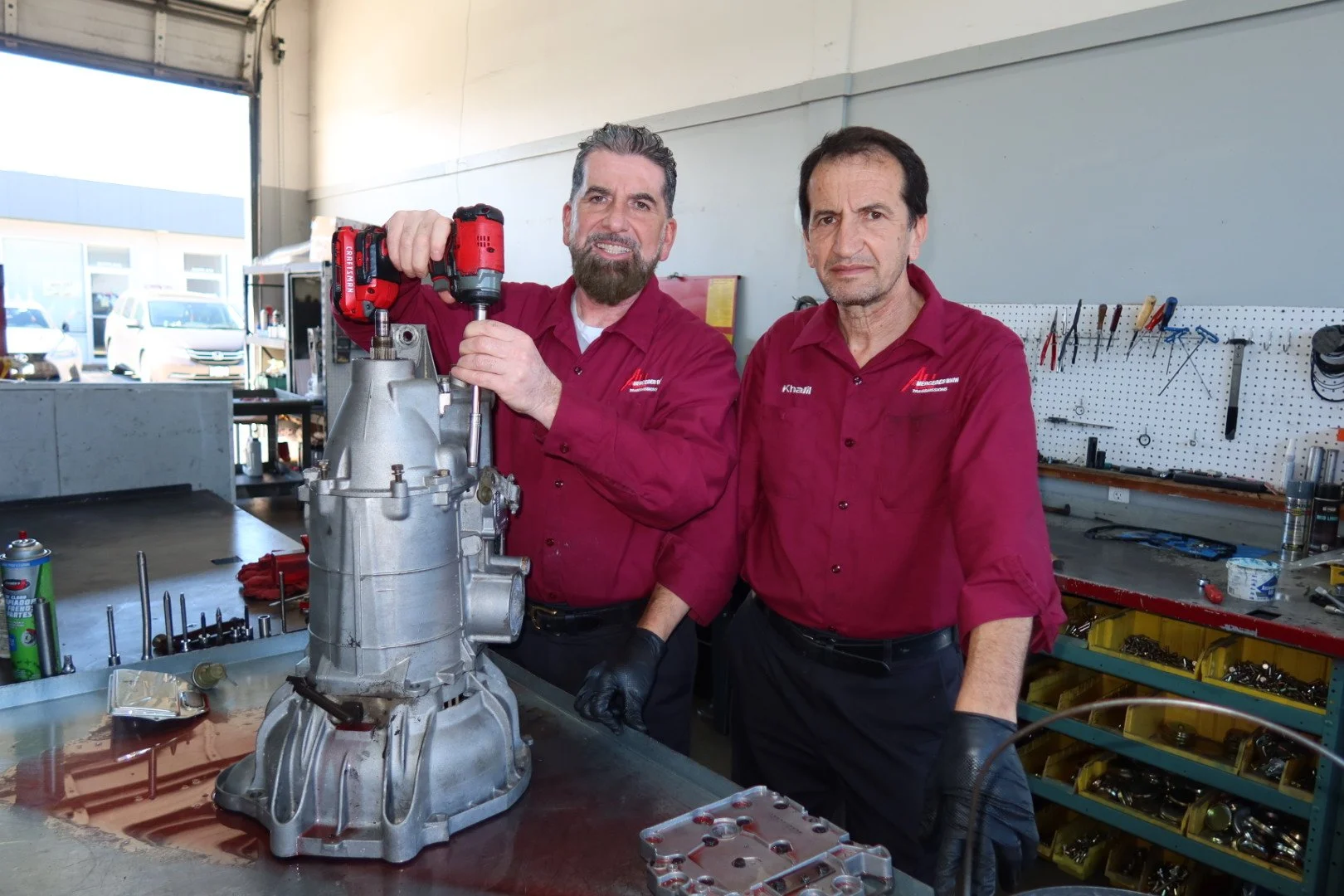 Two men in red uniforms working on a metal car part in a workshop with tools and equipment around.