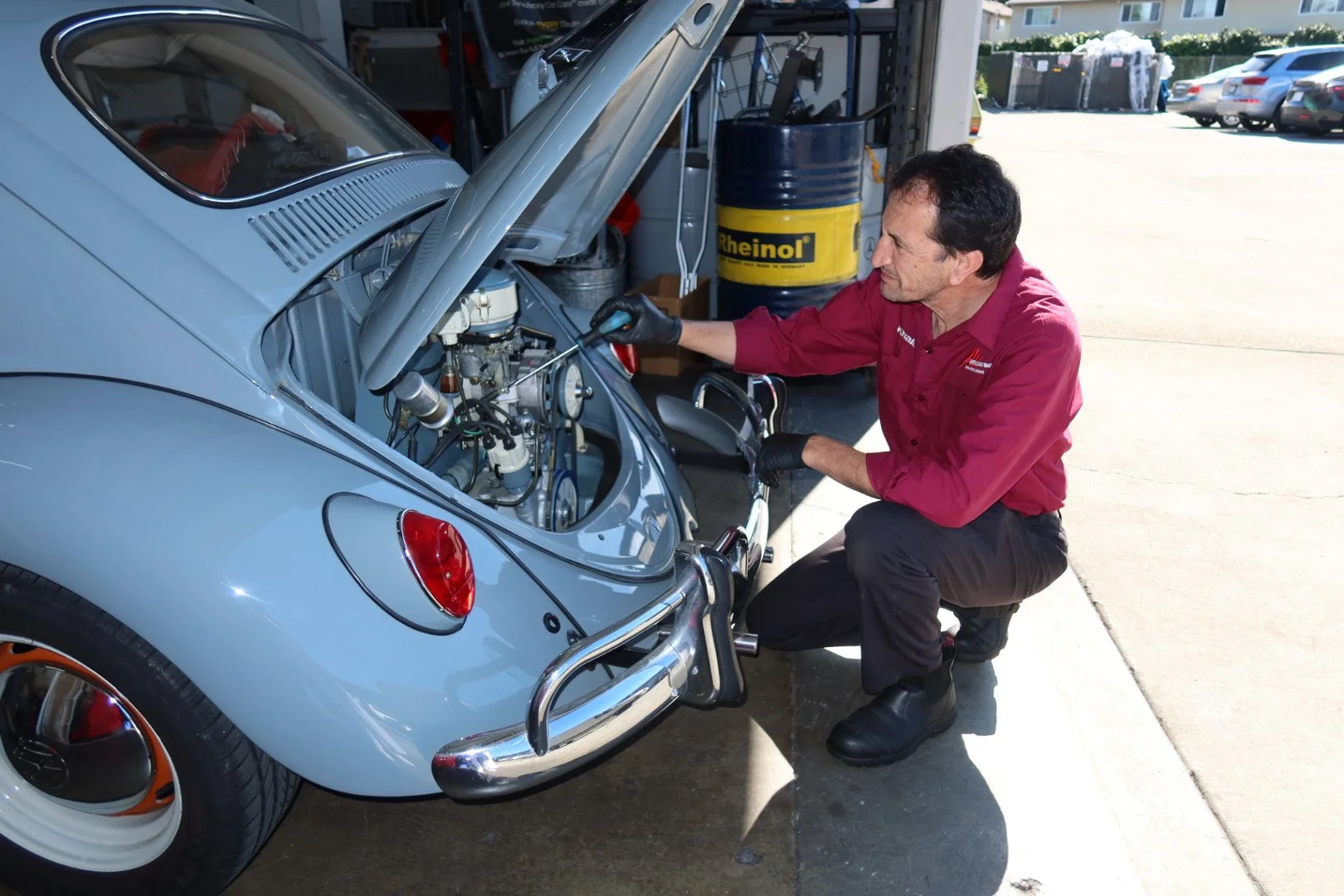 A mechanic in a red shirt and black gloves working on the engine of a classic light blue Volkswagen Beetle in a garage parking lot.