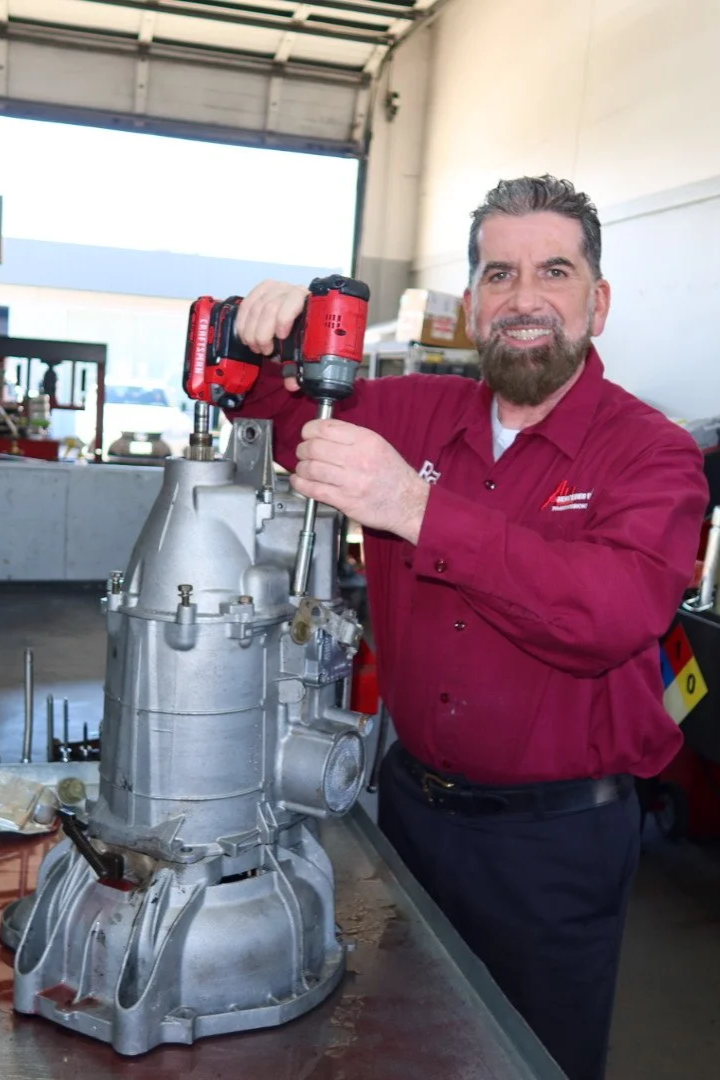 A man in a red shirt is working on a mechanical component using a red cordless power drill inside a garage or workshop.