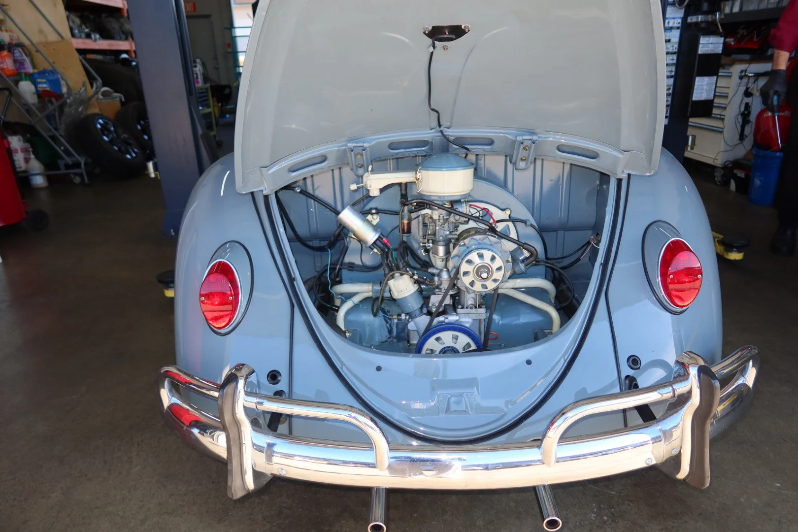 Rear view of a classic Volkswagen Beetle with its engine exposed. The car has a gray exterior, round red tail lights, and a chrome bumper. The orange garage or workshop setting is visible in the background.