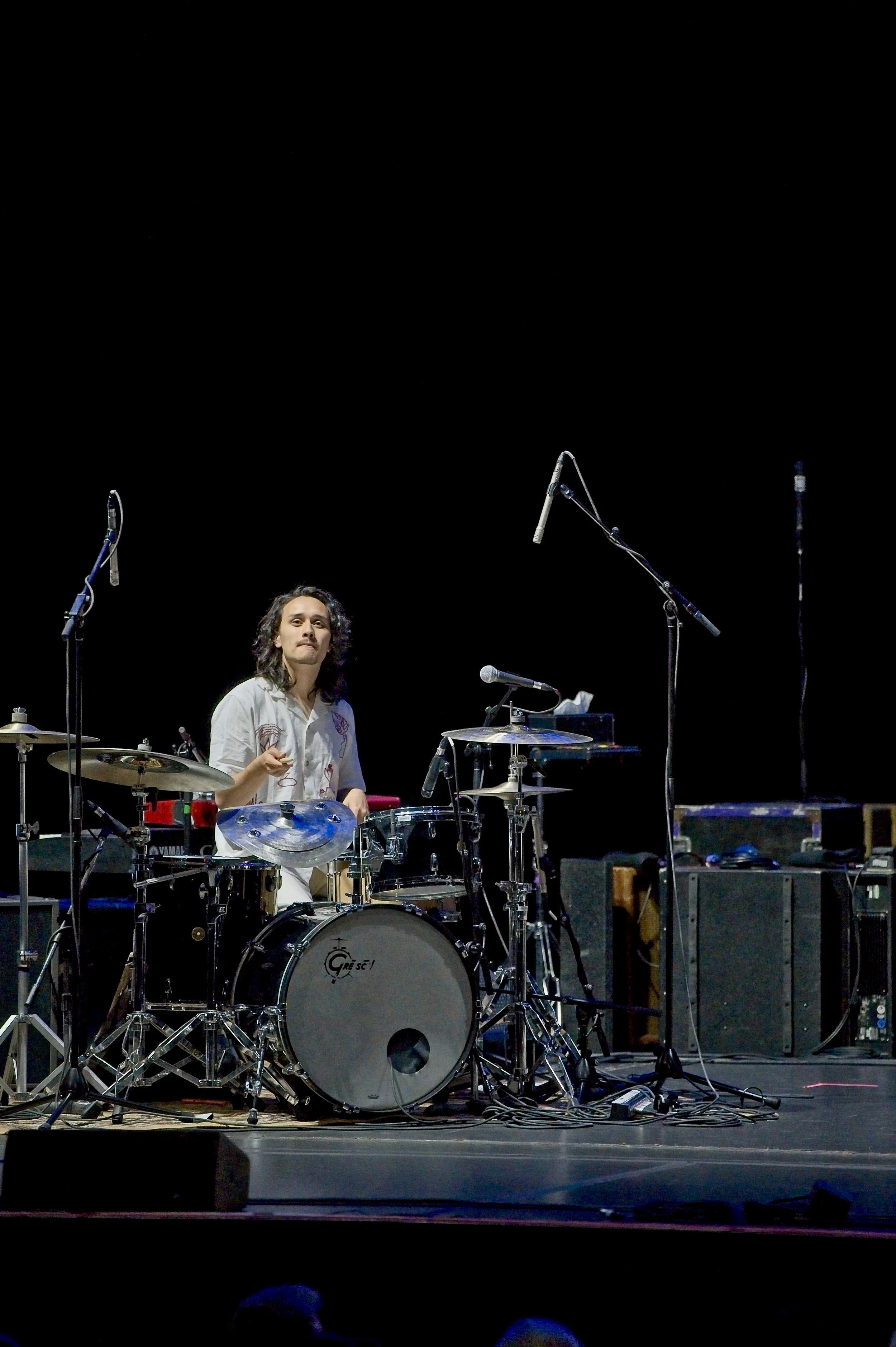 A musician sitting behind a drum set on a stage with a black background, several microphones, and audio equipment.
