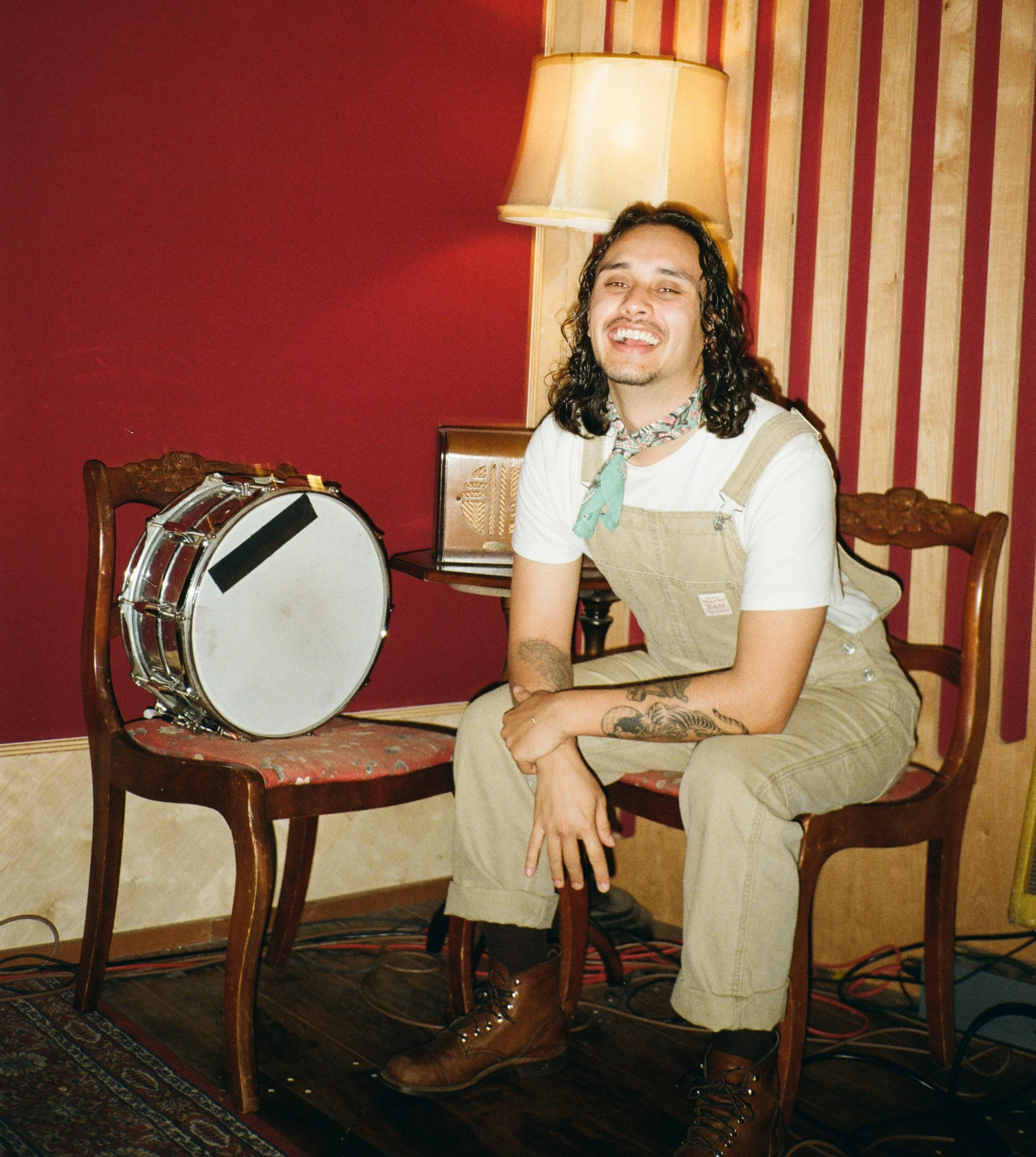 A smiling man with long curly hair and tattoos, seated on a wooden chair in a room with red and striped wallpaper, wearing beige overalls, a white t-shirt, and a colorful neck scarf, next to a snare drum, with a lamp and radio in the background.