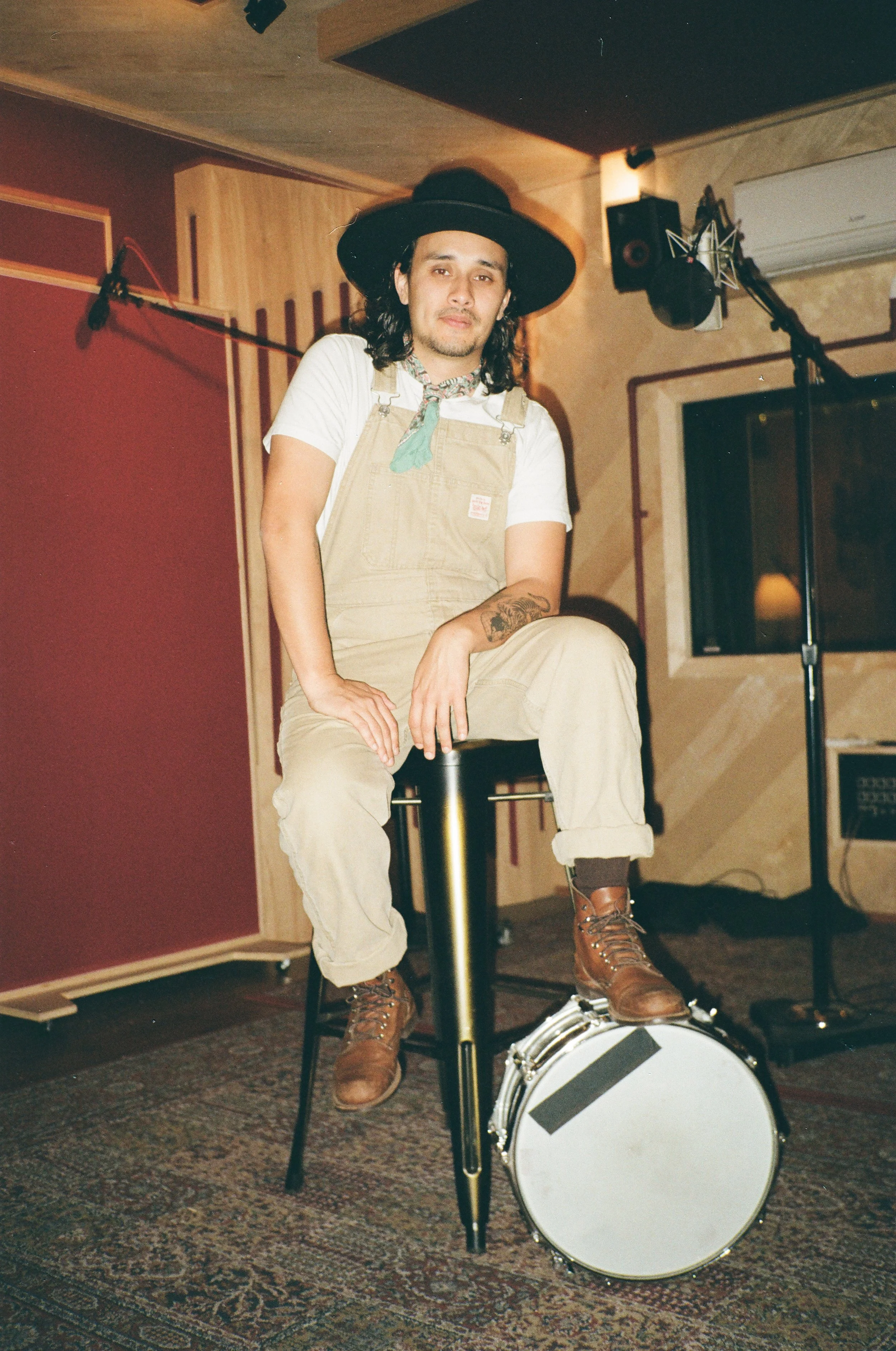 A man in a black wide-brimmed hat and beige overalls sitting on a stool with one foot on a drum in a recording studio.