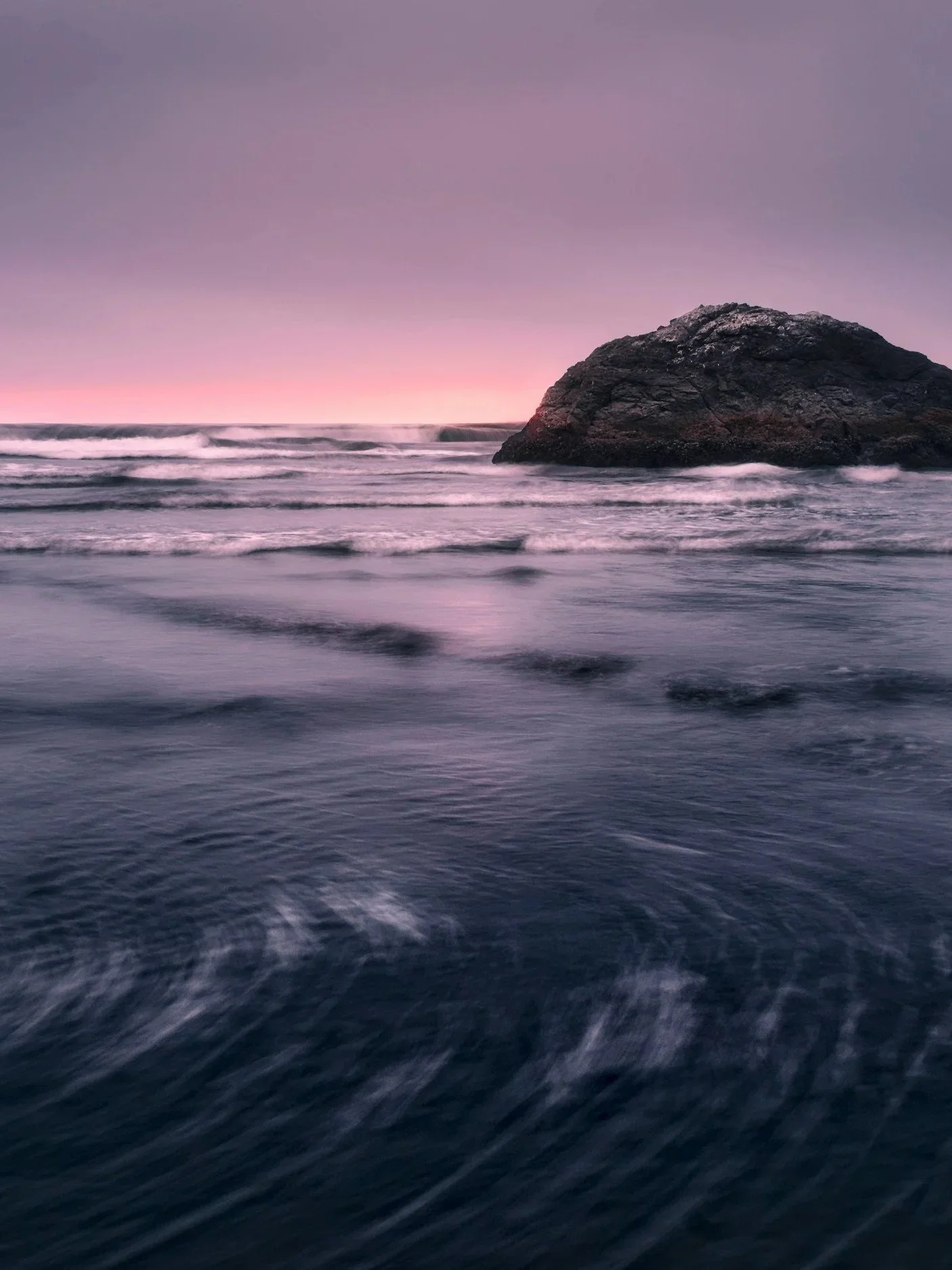 A large rock in the ocean at sunset, with purple and pink sky and waves hitting the shore.