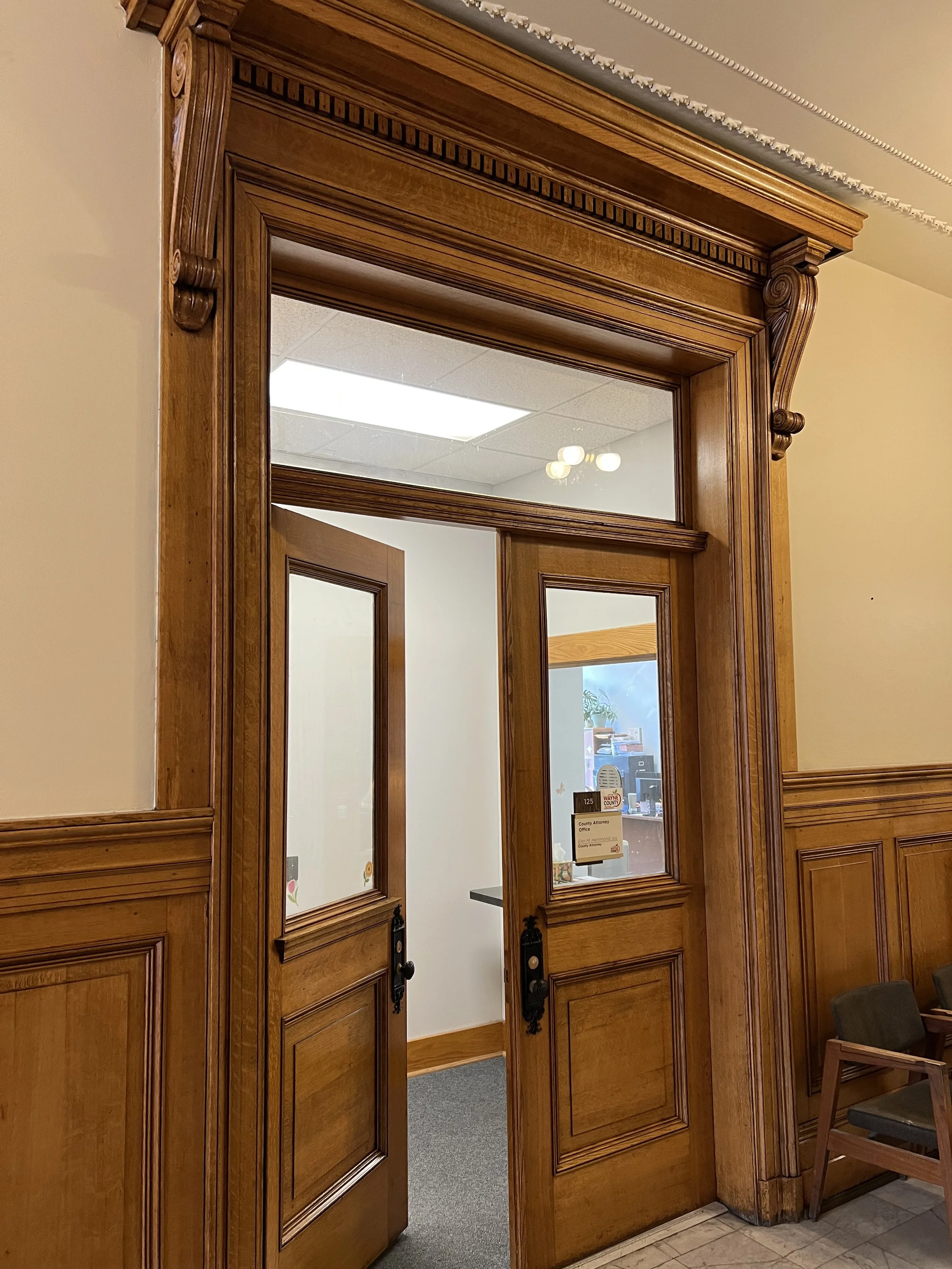 Wooden doorway with a glass window; inside an office with a desk, plant, and computer monitor.