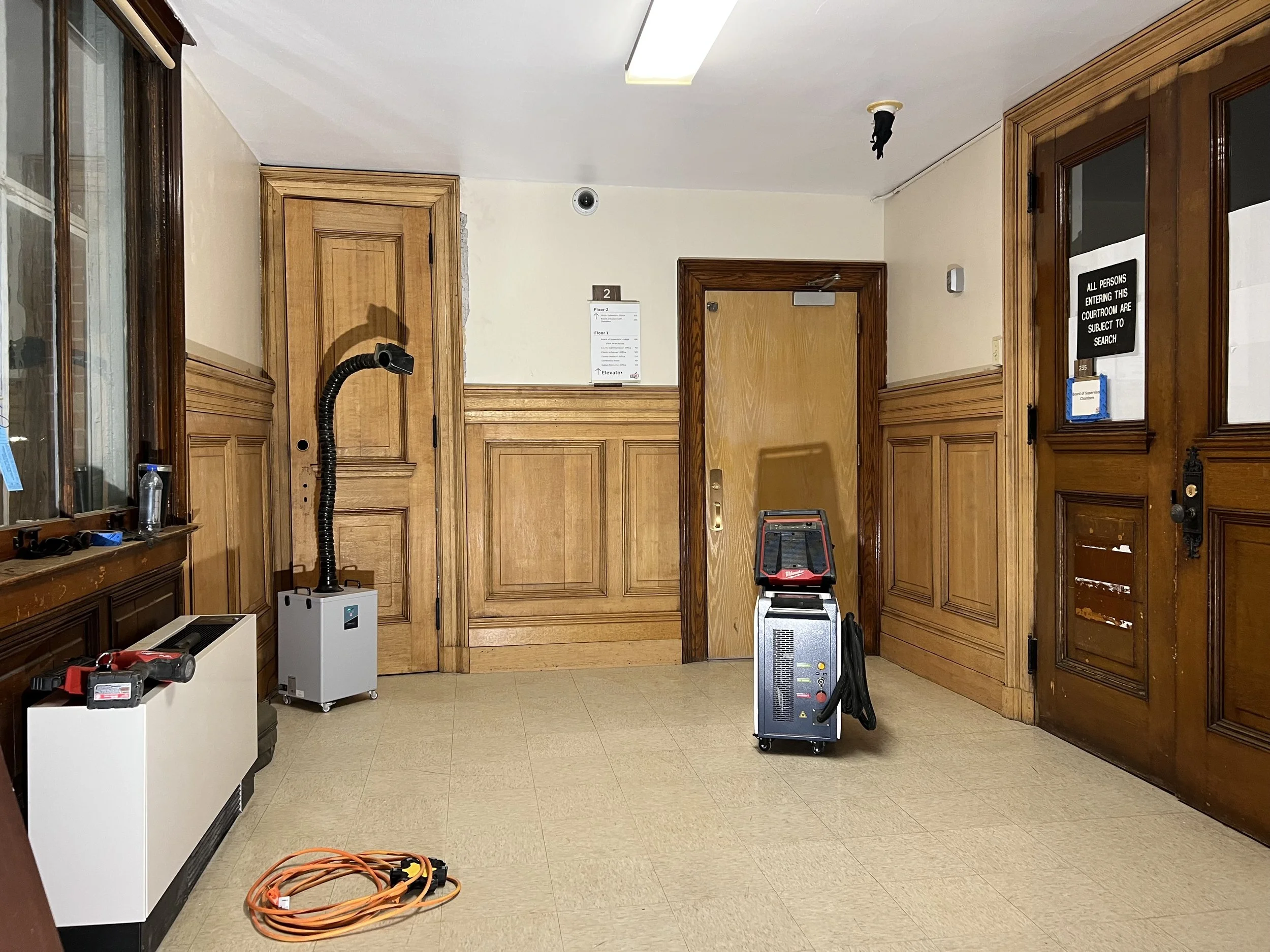 Interior view of a room with wooden doors and panels, a sign on the wall, and equipment such as vacuum, fume extractor, and cords on the floor.
