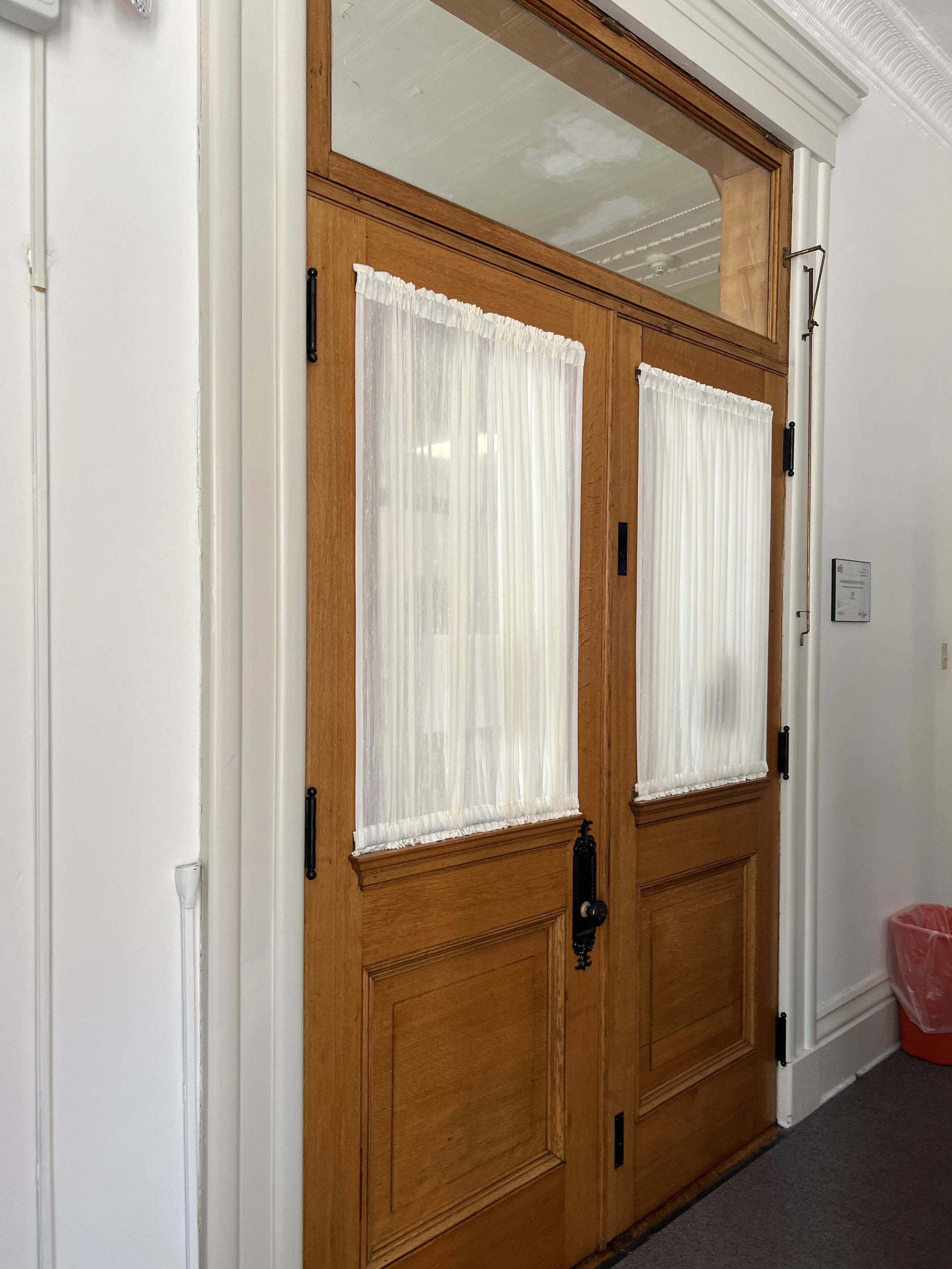 Wooden double door with curtains and a mirror at the top, white walls, a small plaque on the wall, and a red trash can in the corner.