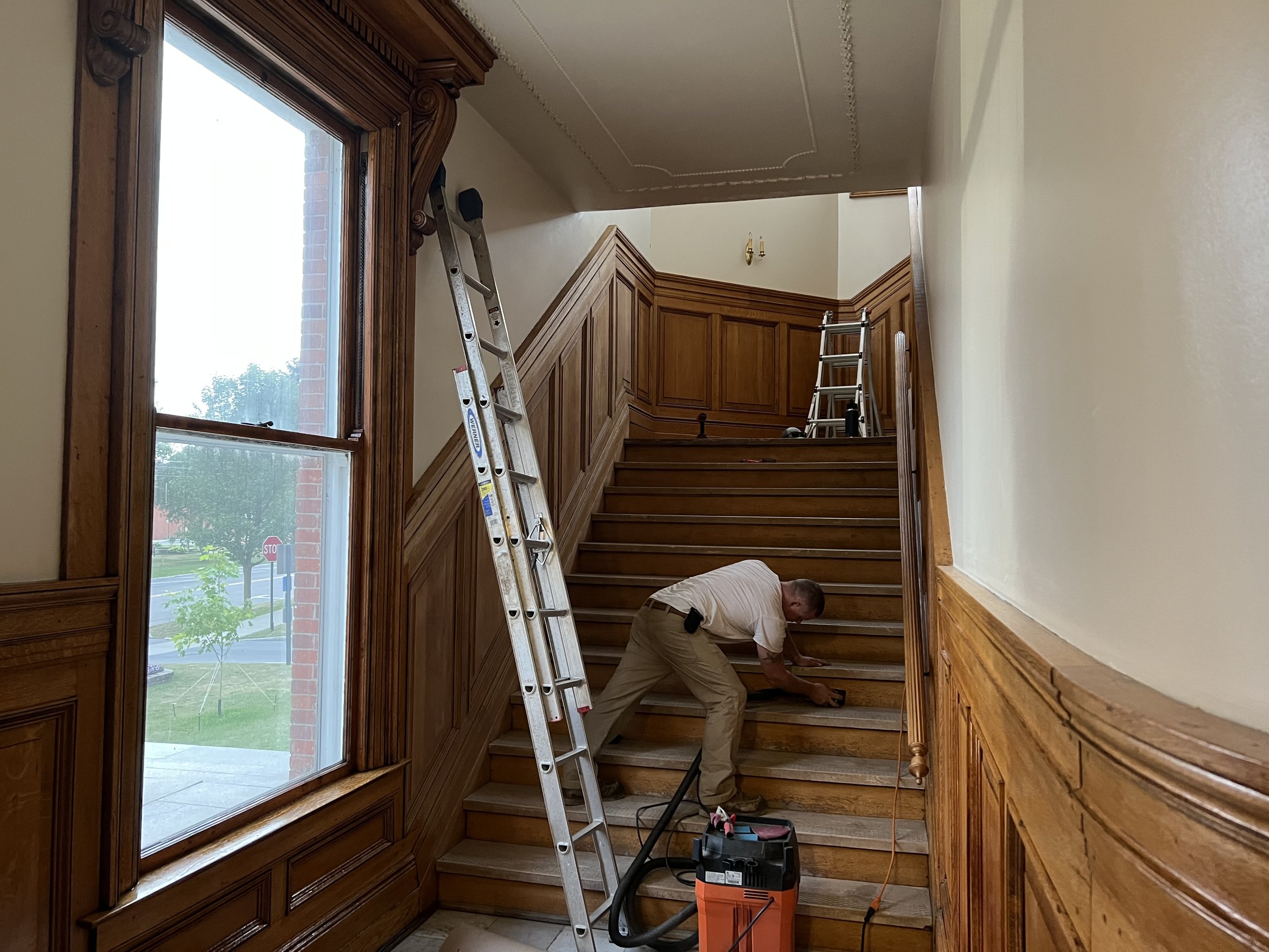 A man working on wooden stairs inside a home, with ladders leaning against the wall, a window to the left, and a vacuum cleaner nearby.