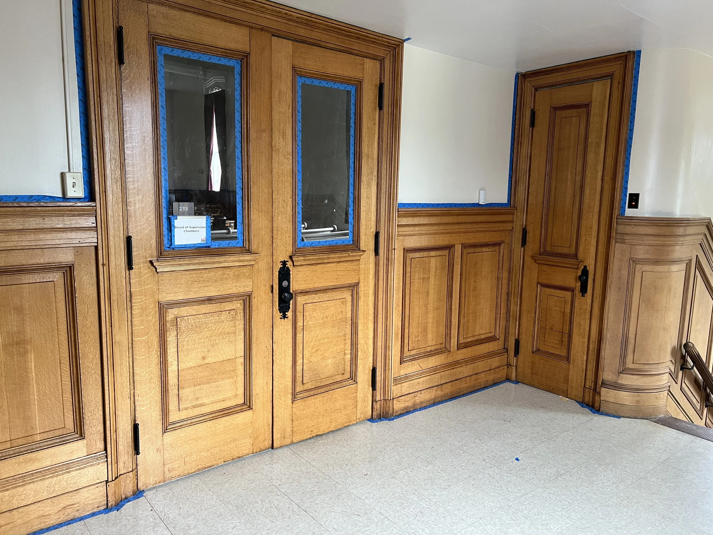 Wooden office partition with doors, painted blue painter's tape along the edges, and a sign that reads 'Board of Supervisor Chambers.' Beige tiled floor and a wall with white paint and a black outlet at the corner.