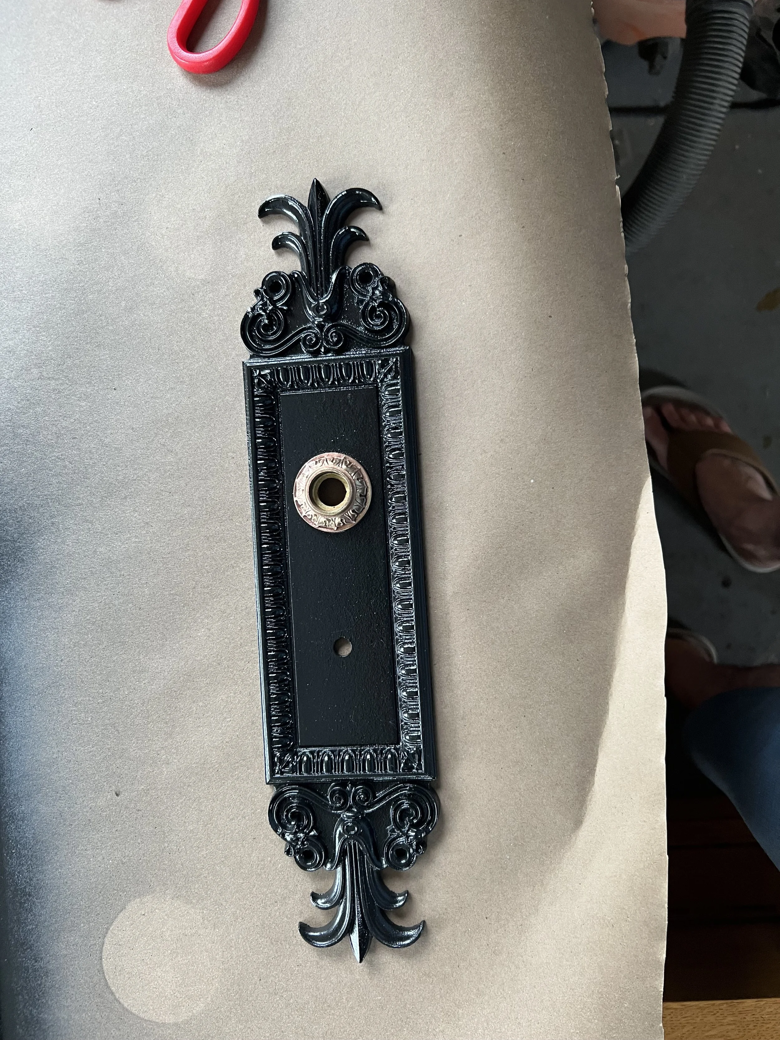 A decorative black metal wall plaque with ornate scrollwork and a rectangular center featuring a circular attachment point.