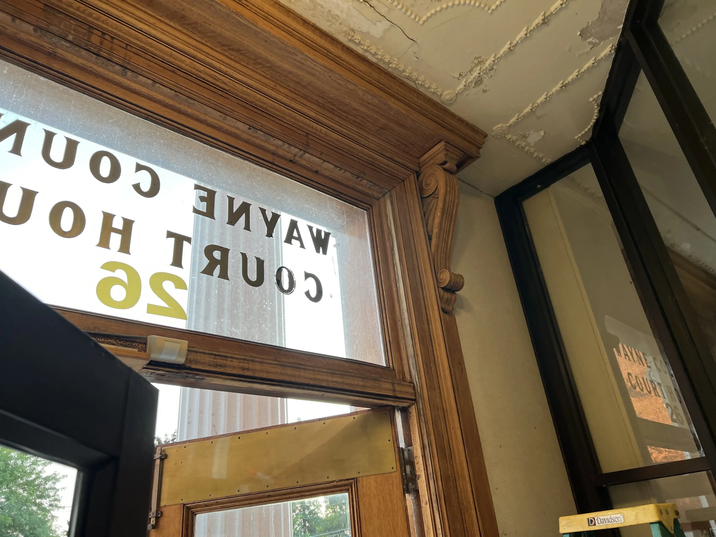 A wooden window frame with glass panes, part of a storefront or office with the words, "WAYAN COURT" and "COU" visible on the window. A small yellow Davidson level is on a shelf or ledge below. The ceiling has peeling paint and decorative molding, and a black metal frame is on the right.
