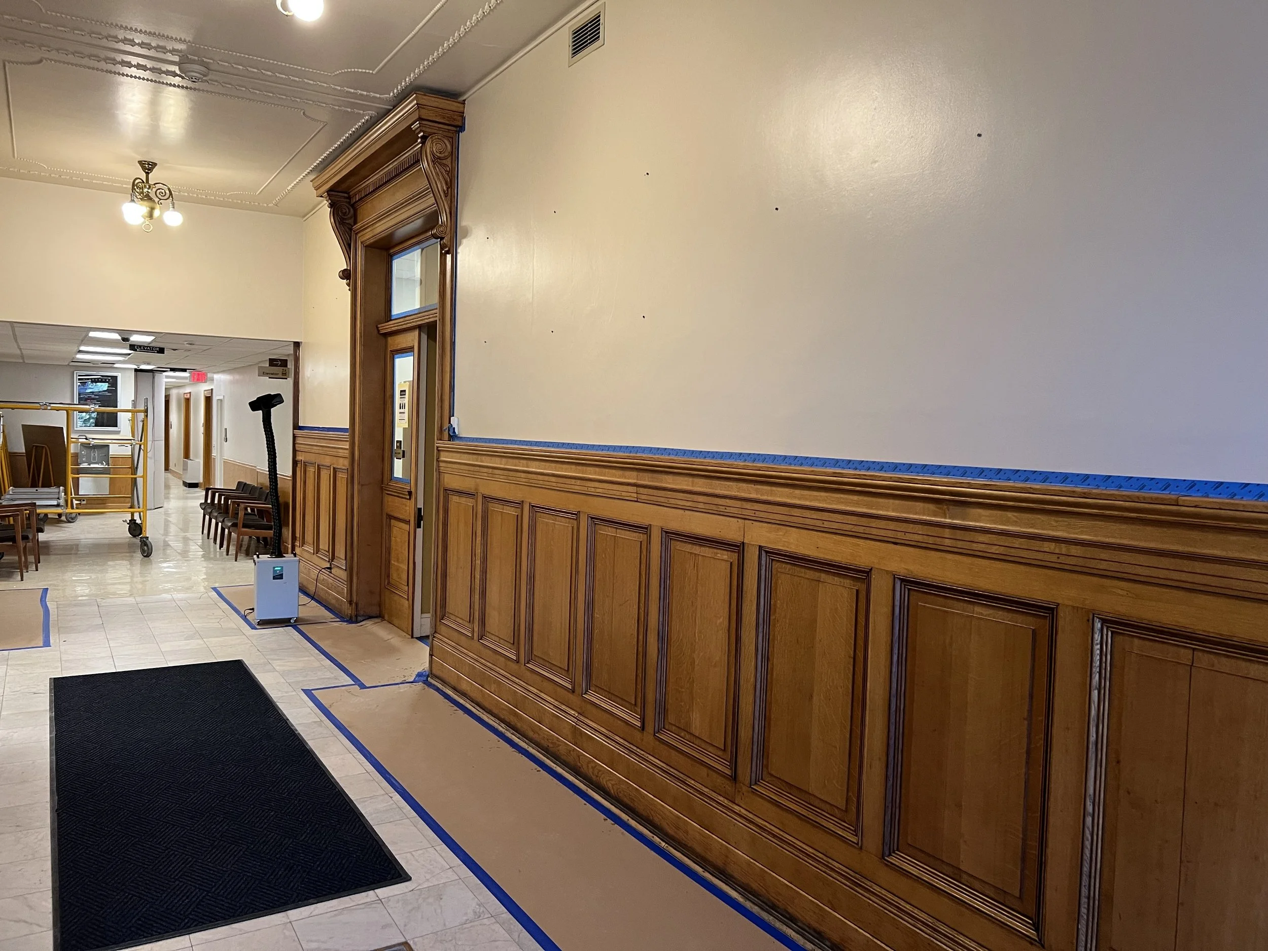 Interior of a building with a wooden wainscot and decorative trim. There are chairs, a yellow rolling cart, and an elevator in the background. Blue painter's tape outlines the base of the wall and some objects on the floor.
