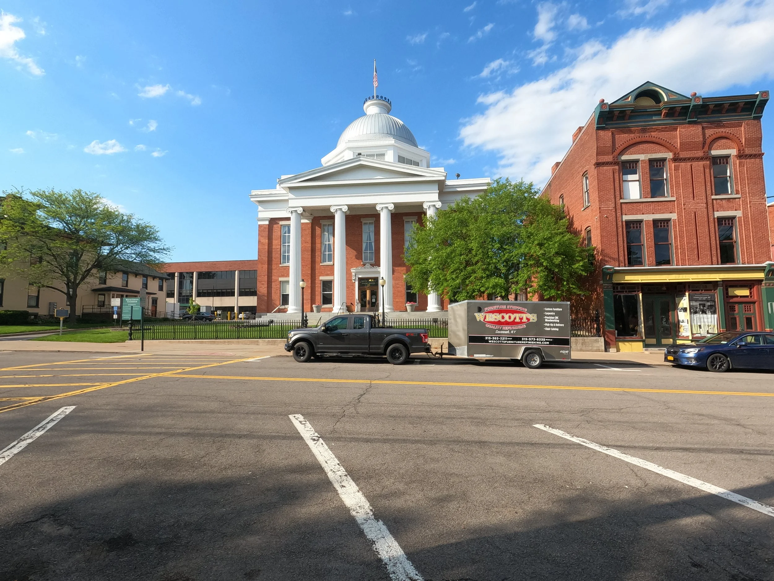 Street view with a historic courthouse building featuring a white dome and large columns, situated between trees and older brick buildings, with parked cars and a our rig ready to unload in the foreground.