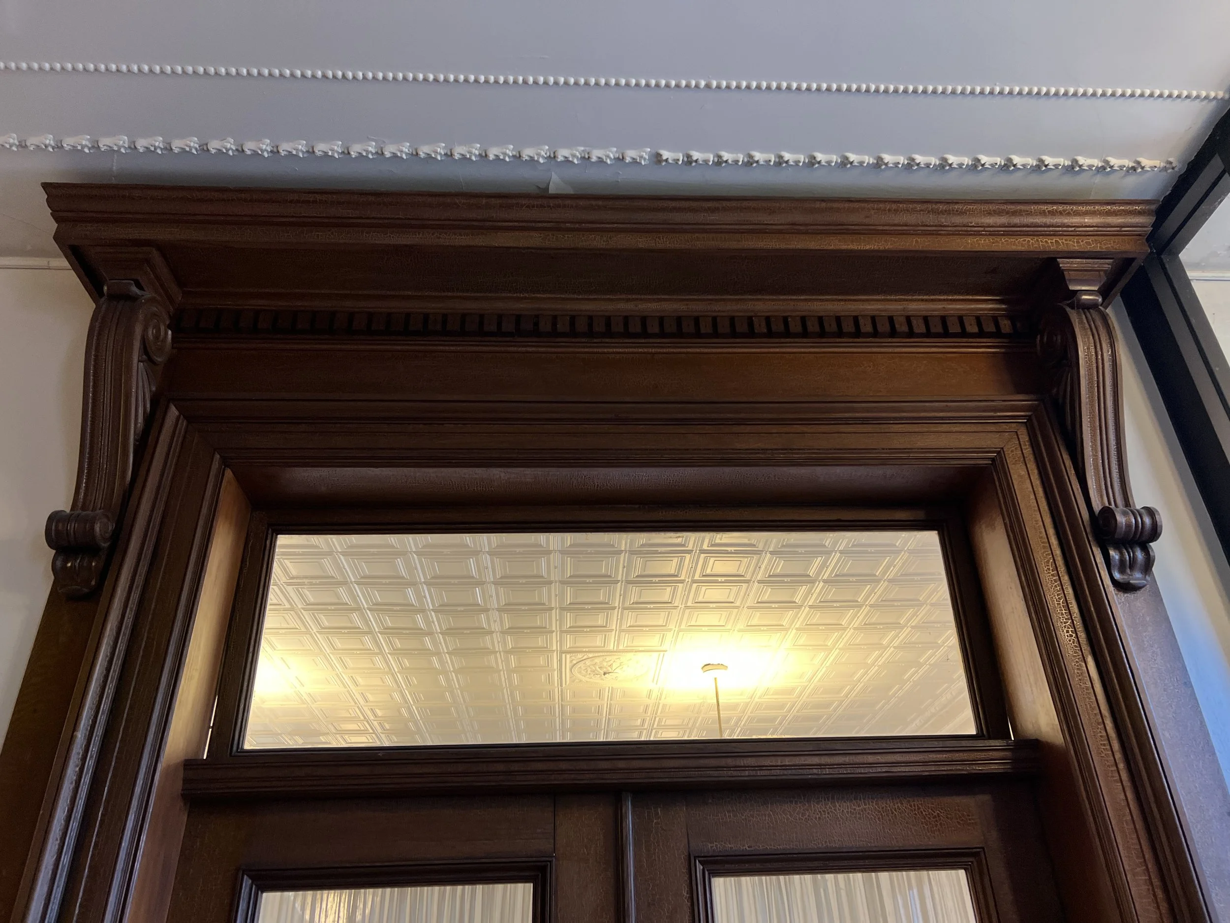 Interior view of a room with a large wooden doorway, ornate wood trim, and a ceiling with patterned tiles and a hanging light fixture.