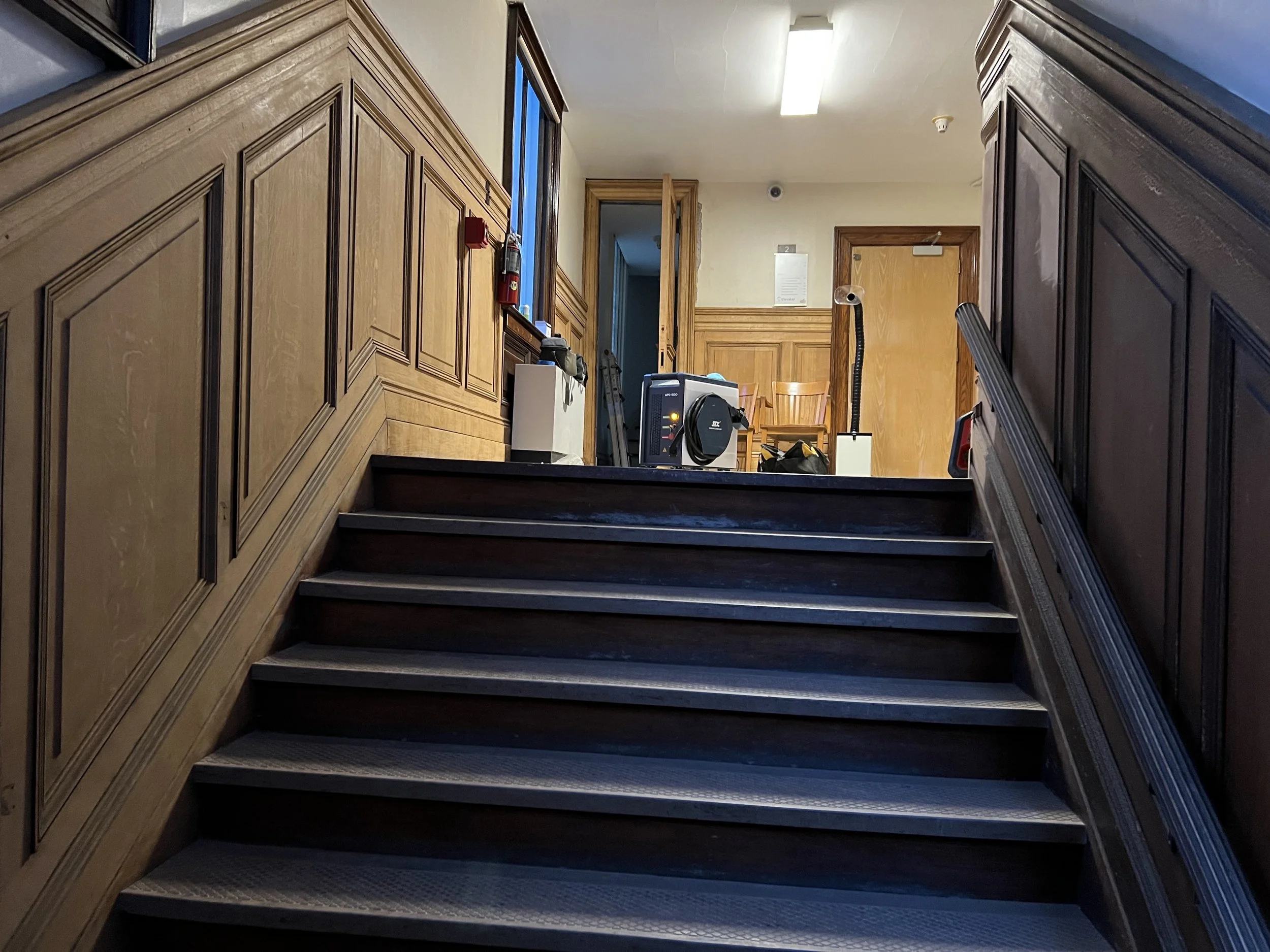 An indoor staircase with dark steps and wood-paneled walls on either side, leading up to a landing with chairs and miscellaneous equipment.