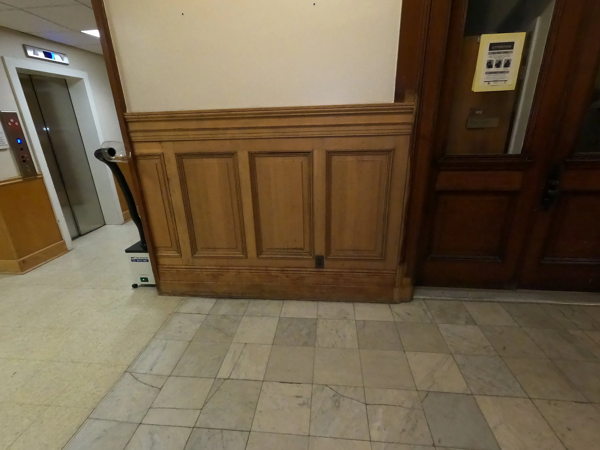 Inside an elevator lobby corridor with an elevator door on the left, a decorative wooden wall recrently stripped panel middle, and a mirrored panel with a yellow notice on the right. The floor has beige and grayish tiles.