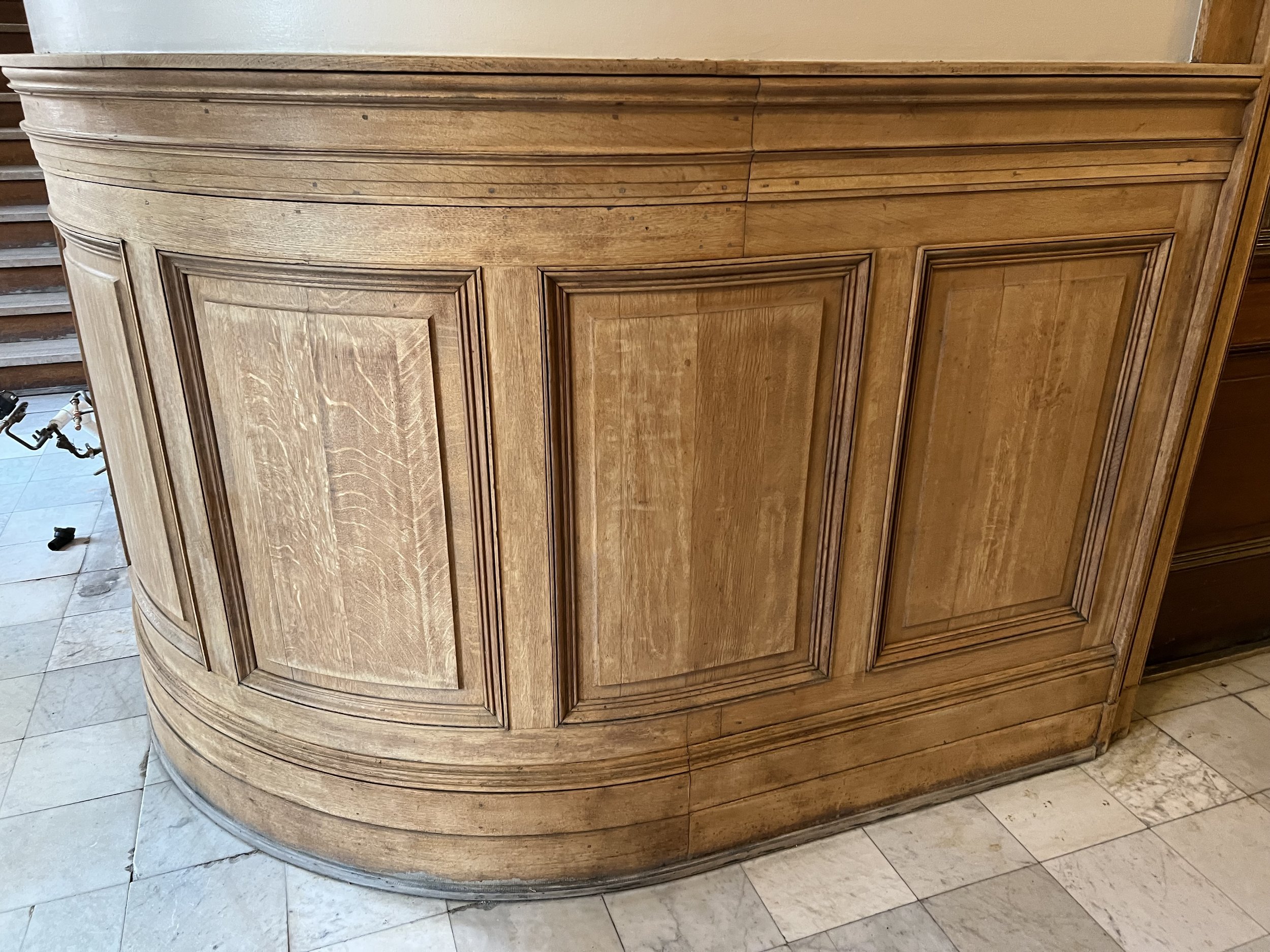 A wooden furniture piece with curved edges, panel detailing, and a natural finish, placed on a tiled floor.
