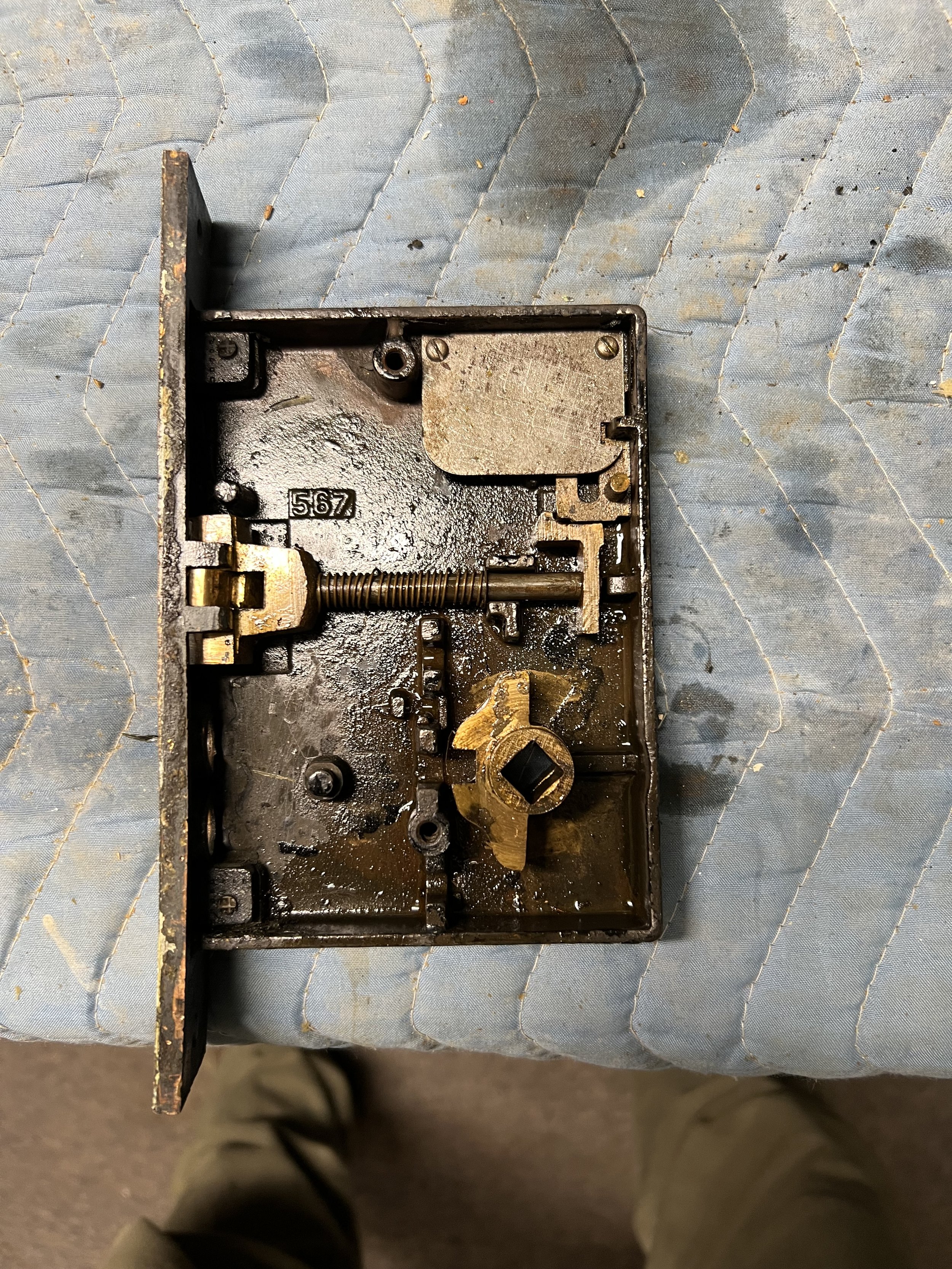 The image shows the inner mechanism of an old mechanical lock, with visible metal parts, springs, and screws, placed on a textured surface.