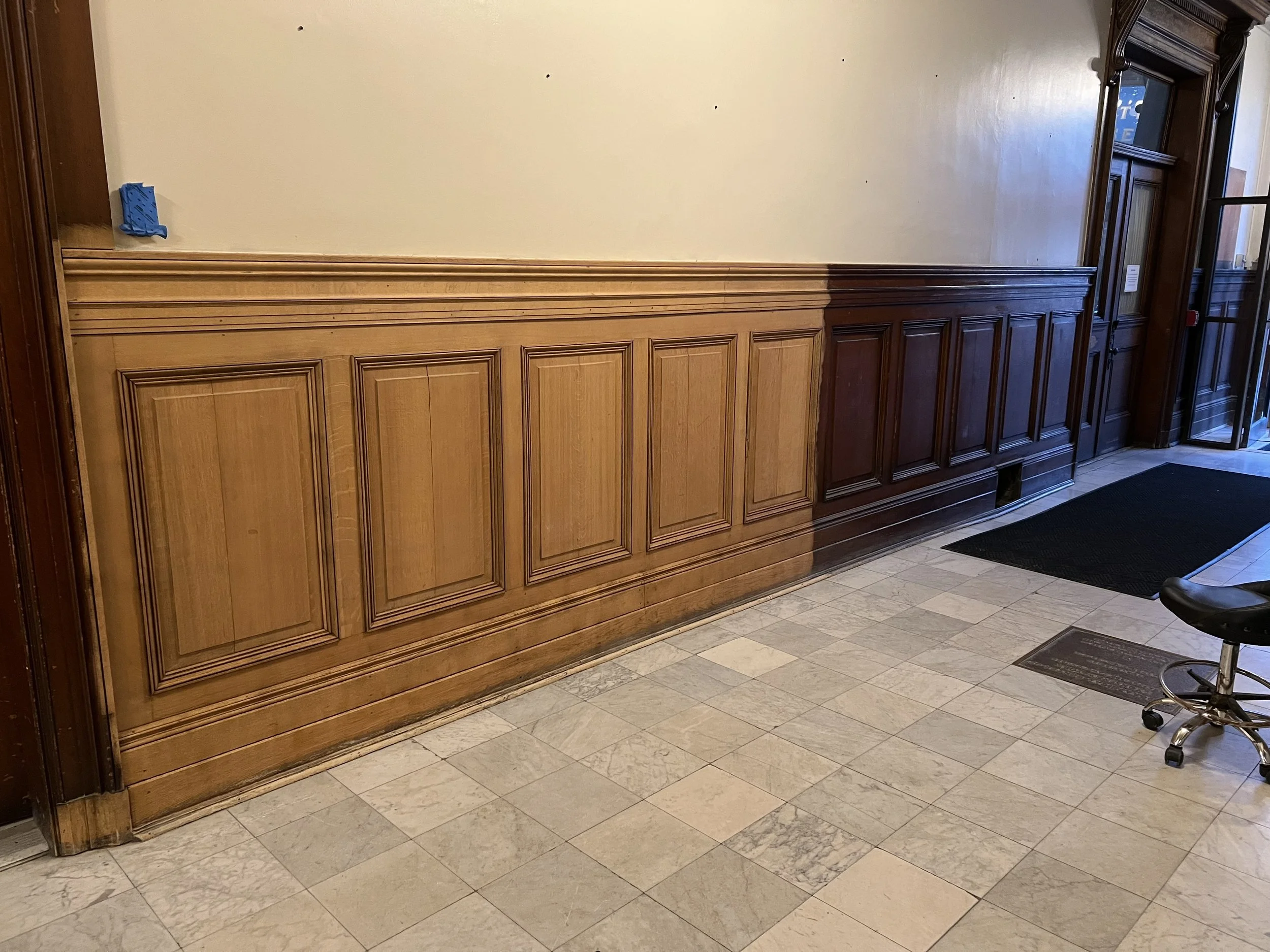 Wood-paneled wall in a building interior with laser stripped panels on left unstripped on right, a black chair with wheels on the right, and a hallway with a door and a window on the far right.
