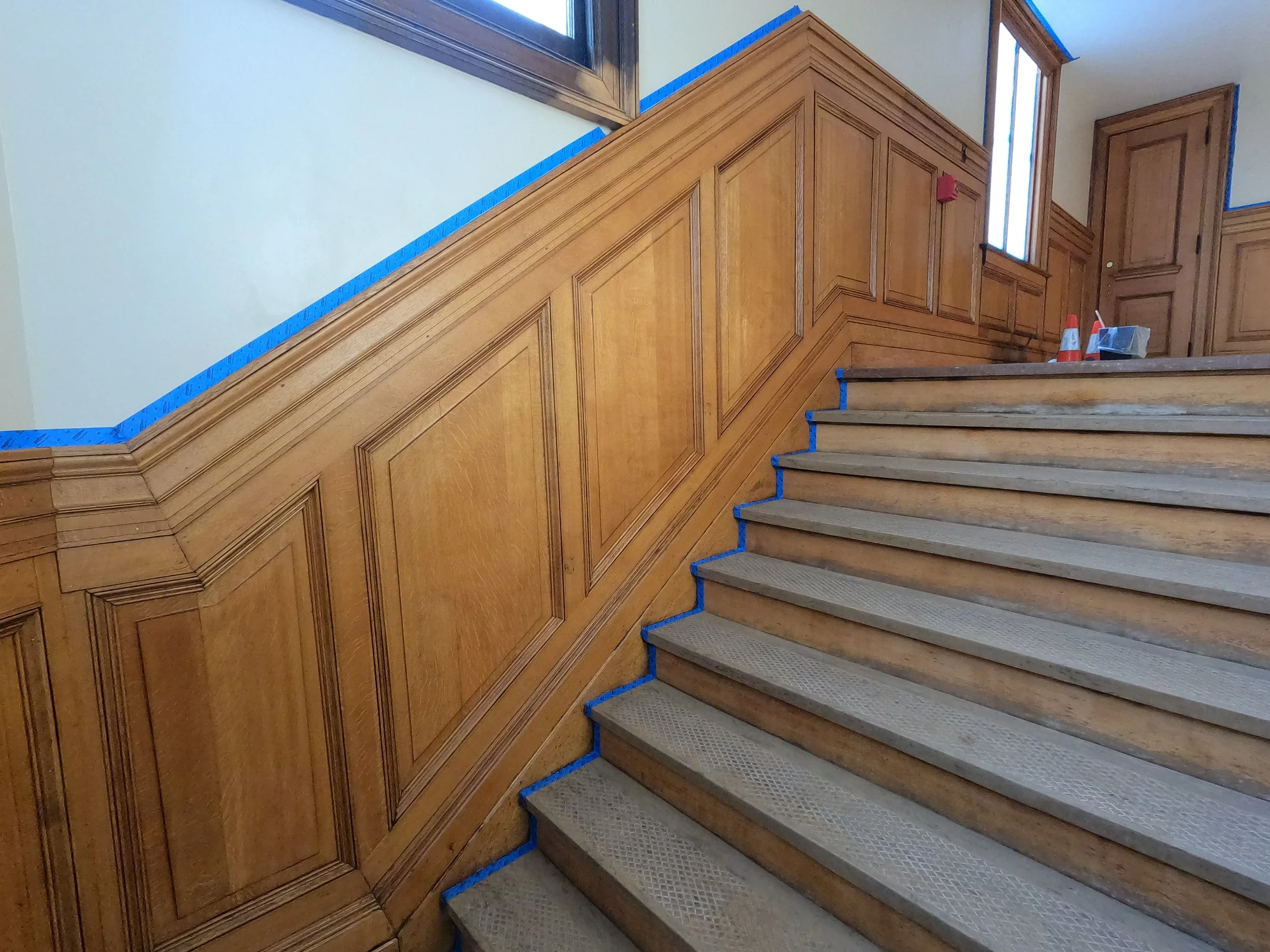 Wooden staircase with a decorative wooden wall panel on the left and a window on the right, with blue painter's tape along the edges of the wall and stairs, and some construction items on the stairs.