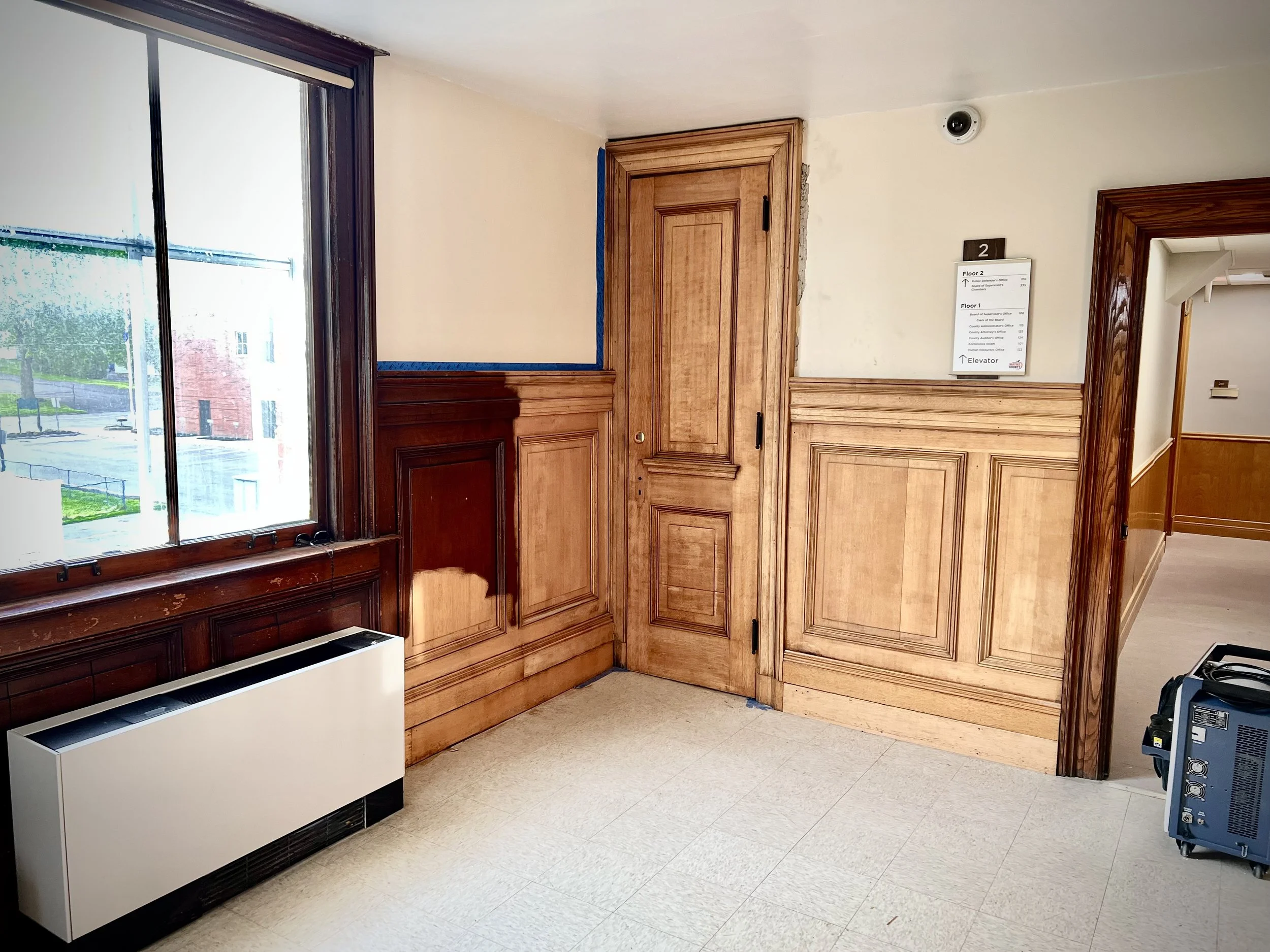 Interior of a room with wood-paneled walls partialy stripped, a wooden door, a large window, and a white radiator . The floor is covered with beige tiles.