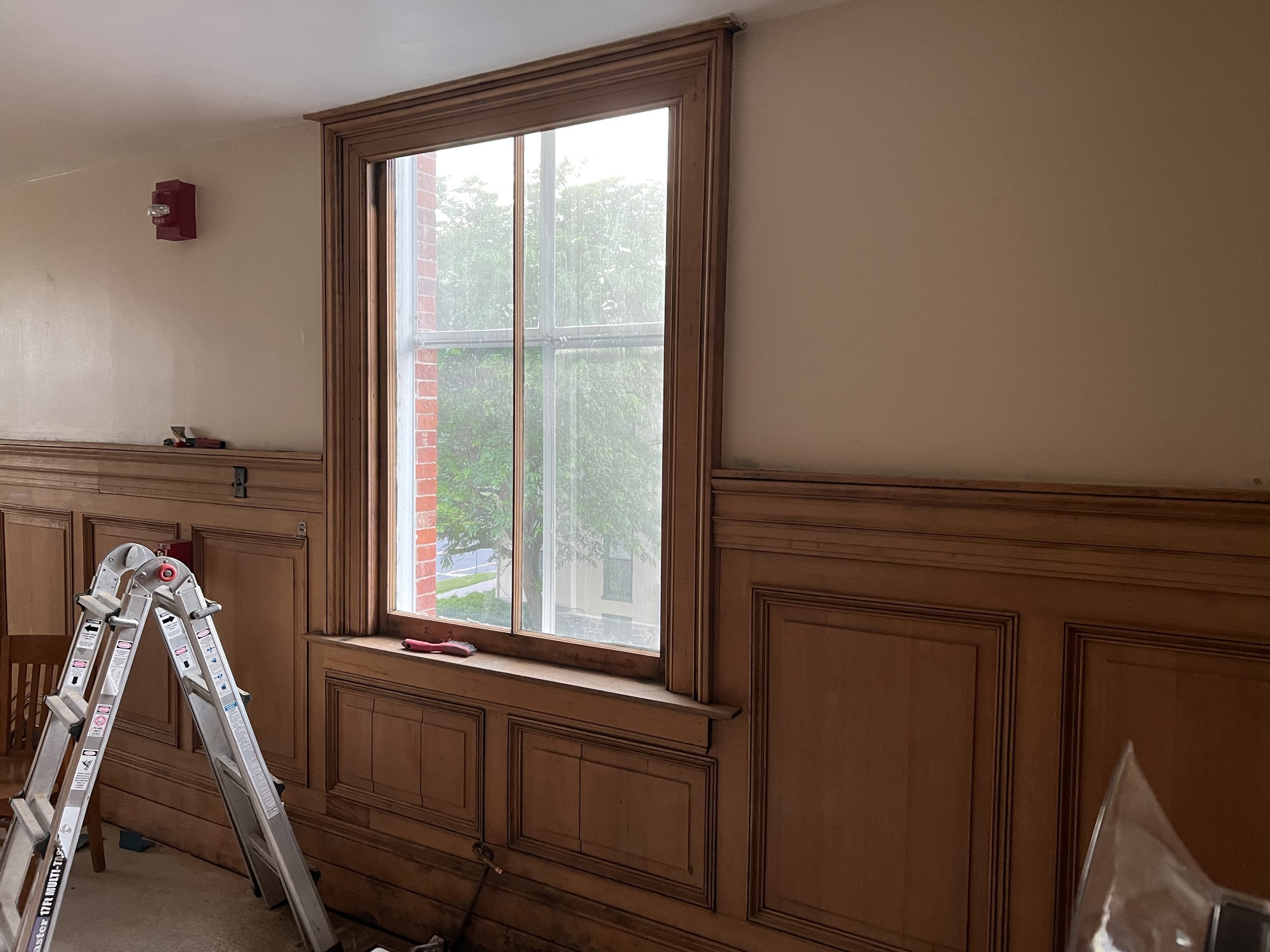 Interior of a room with wooden wall paneling, a large window with a wooden frame, a step ladder, and construction tools, indicating ongoing renovation or construction work.