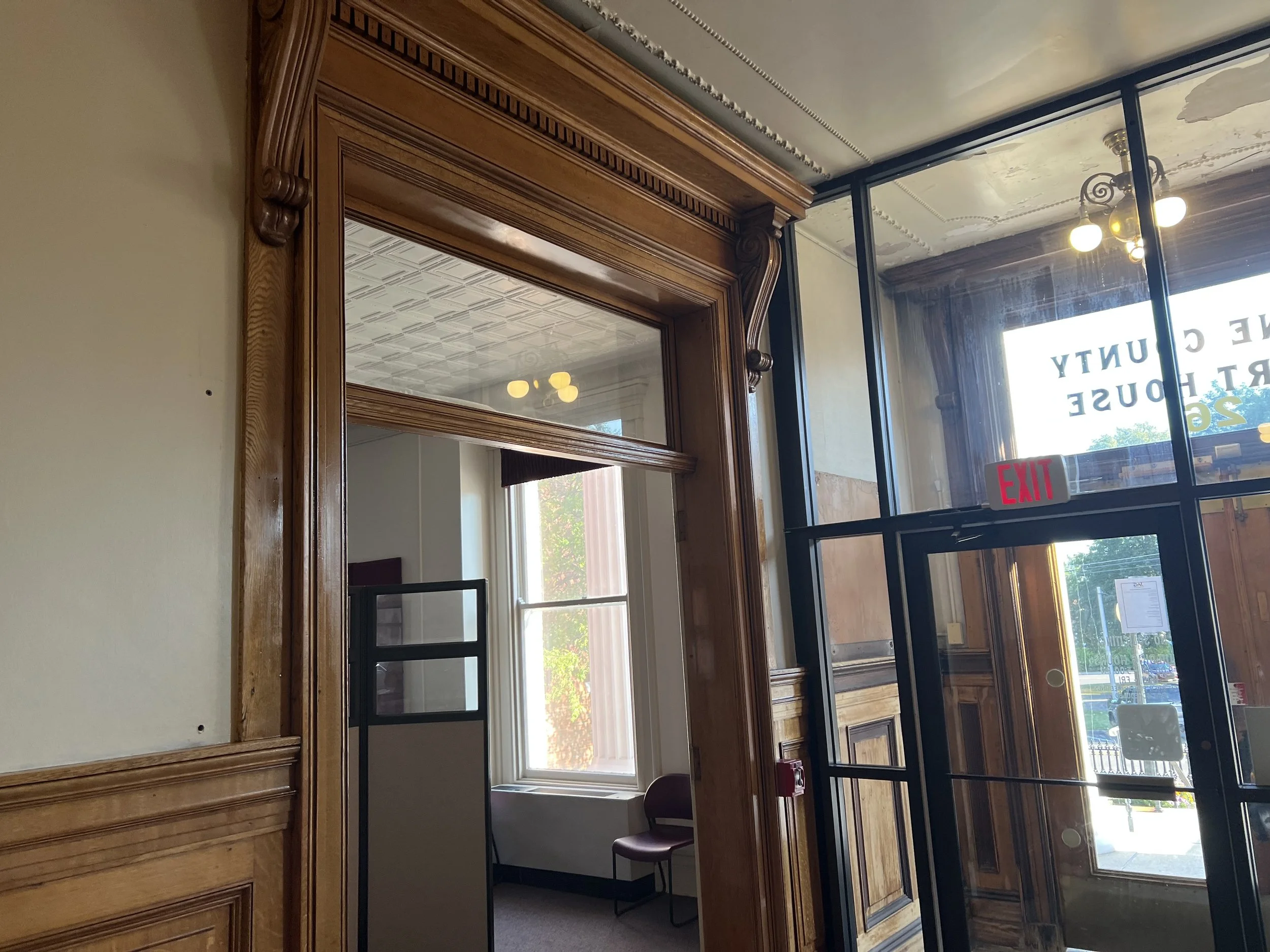 Interior of a building with wooden trim and large glass windows, a ceiling with ornate molding, and a view outside showing trees and a parking lot.