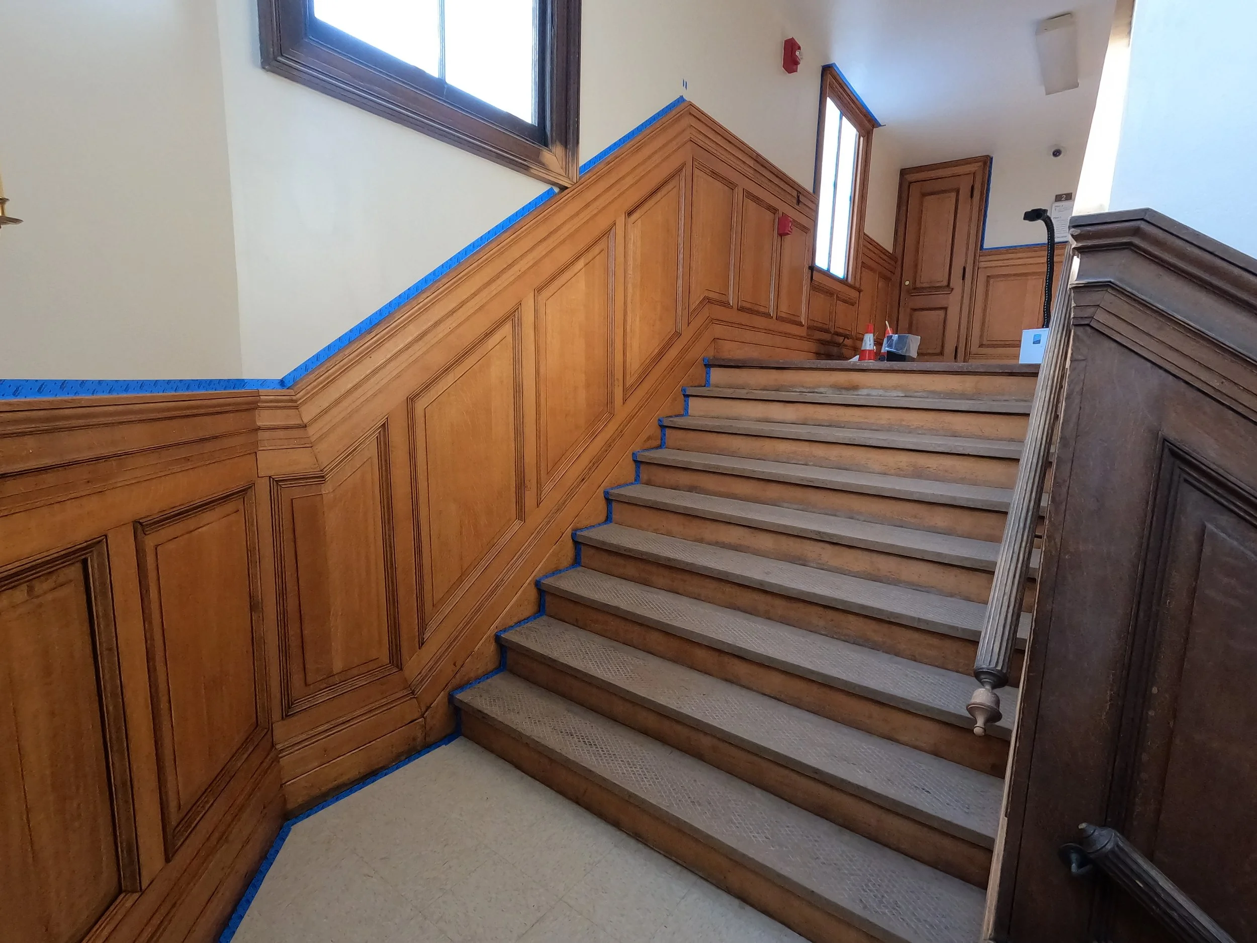 Wood-paneled staircase with carpeted steps and a black handrail, blue painter's tape along the edges of the paneling, and a window at the top of the stairs.
