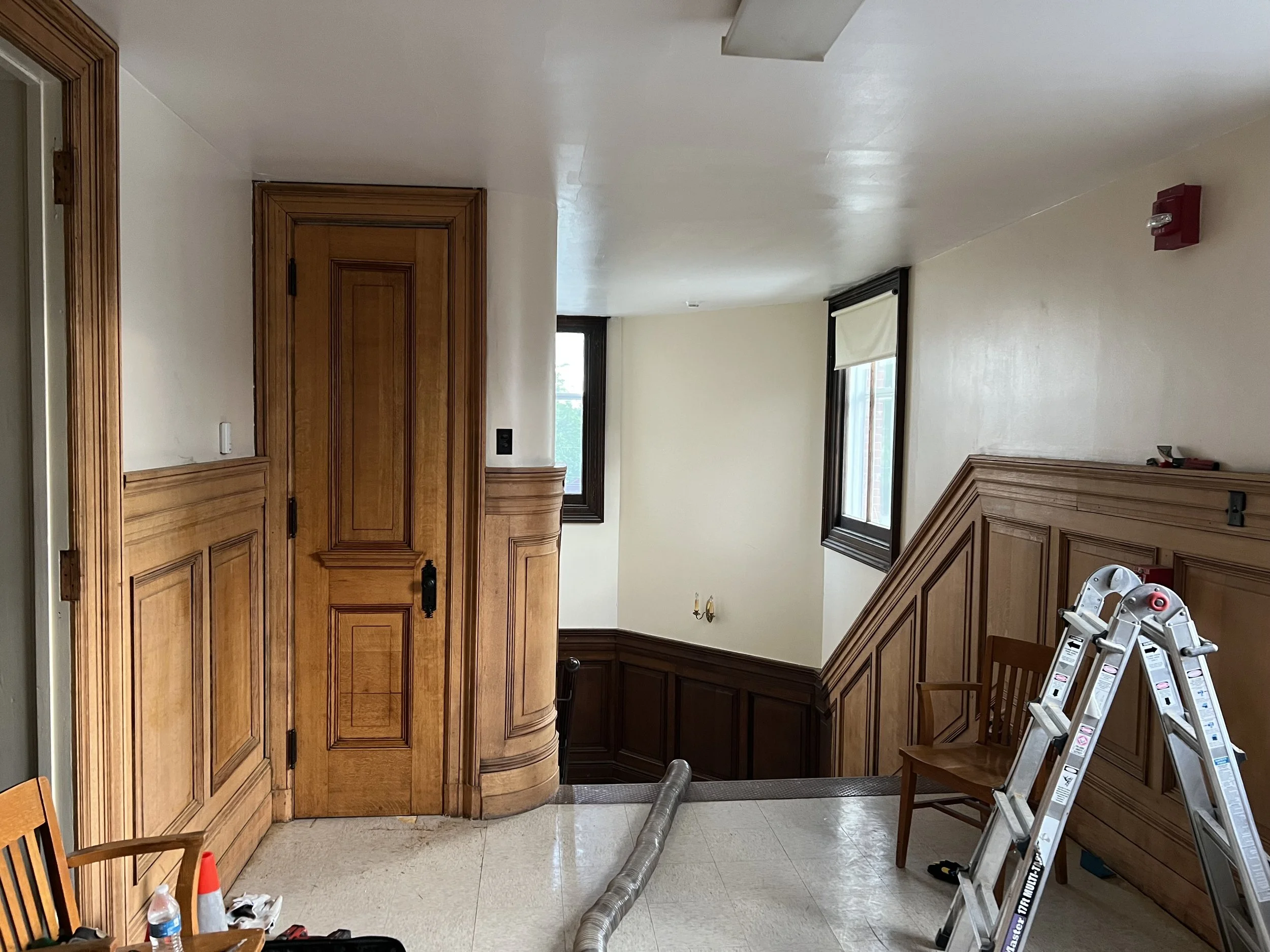 An interior space under construction or renovation with wooden wall paneling, windows, a chair, and a ladder.