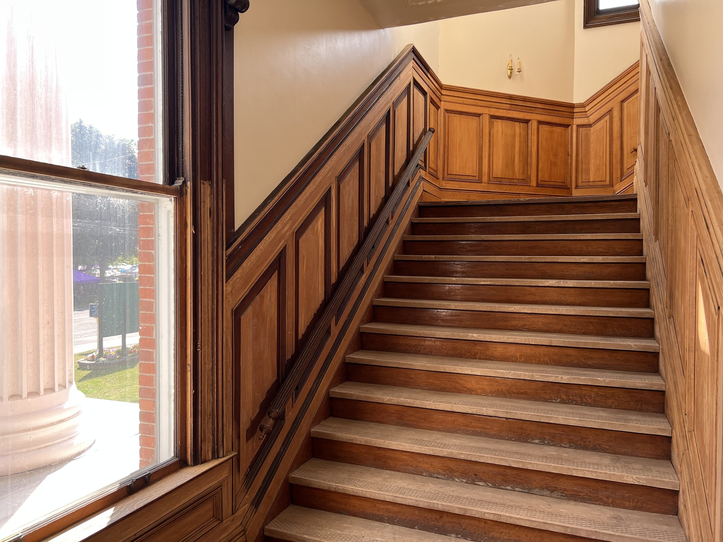 Wooden staircase inside a building, next to a large window with sunlight coming through, and a view of the street outside.