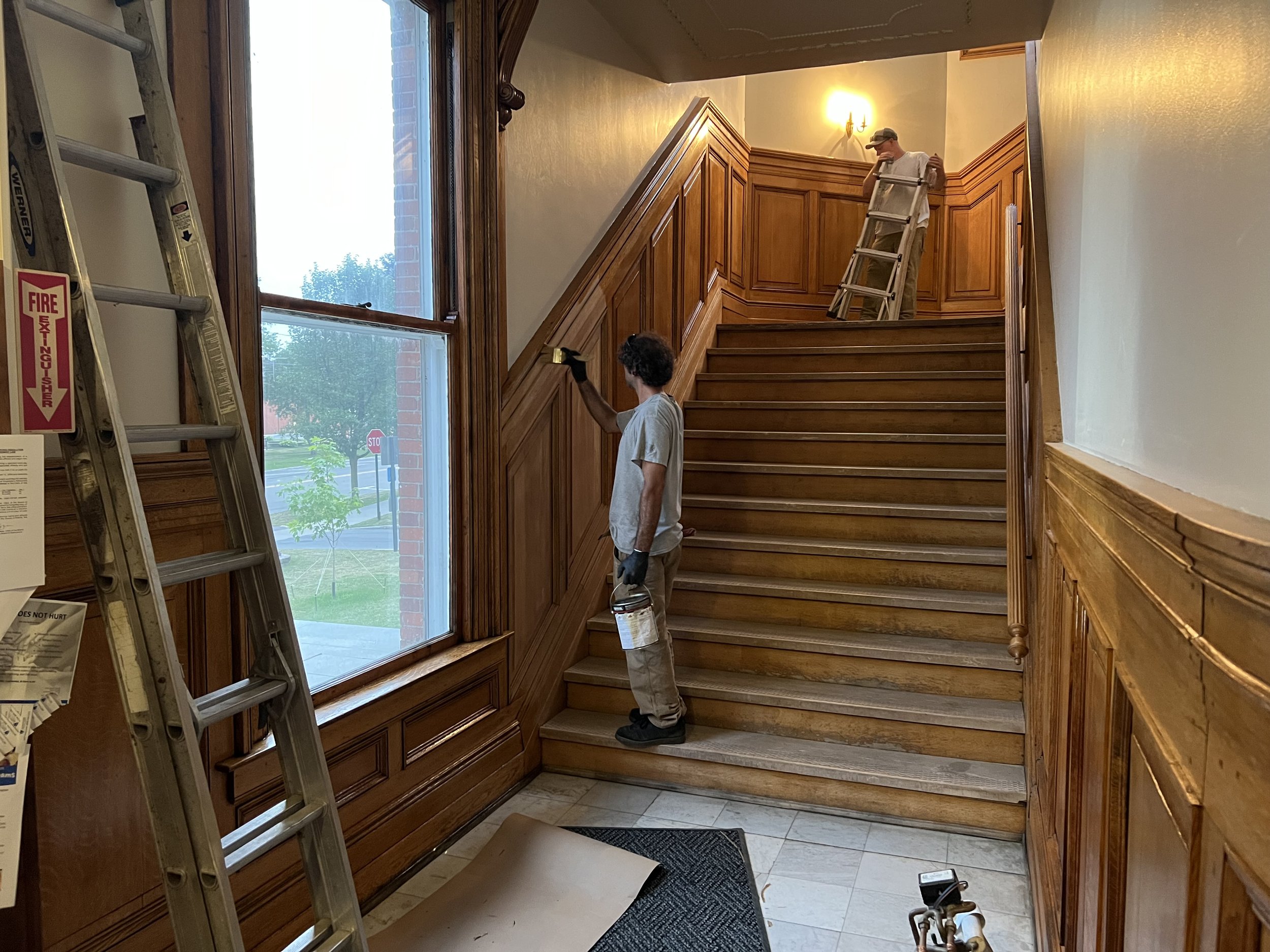 Two workers installing or repairing wooden stairs and wall paneling inside a building, with one climbing a ladder and the other standing on the stairs holding a tool.