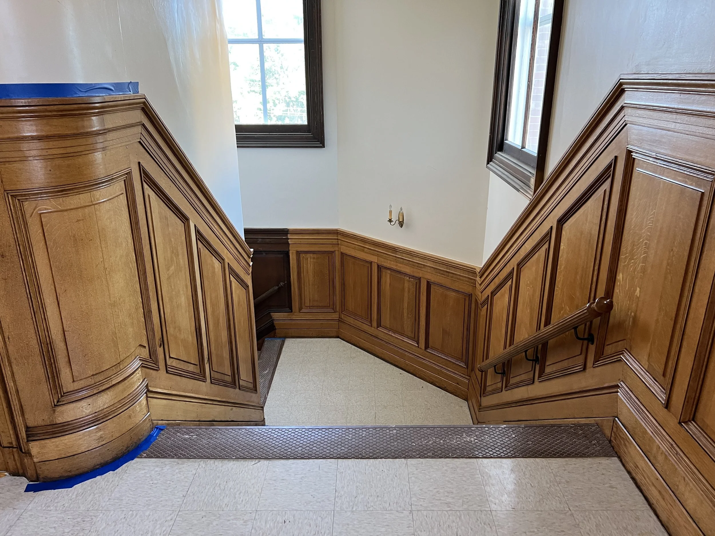 Indoor staircase with wooden paneling, two large windows, and a chandelier fixture on the wall at the top of the stairs.
