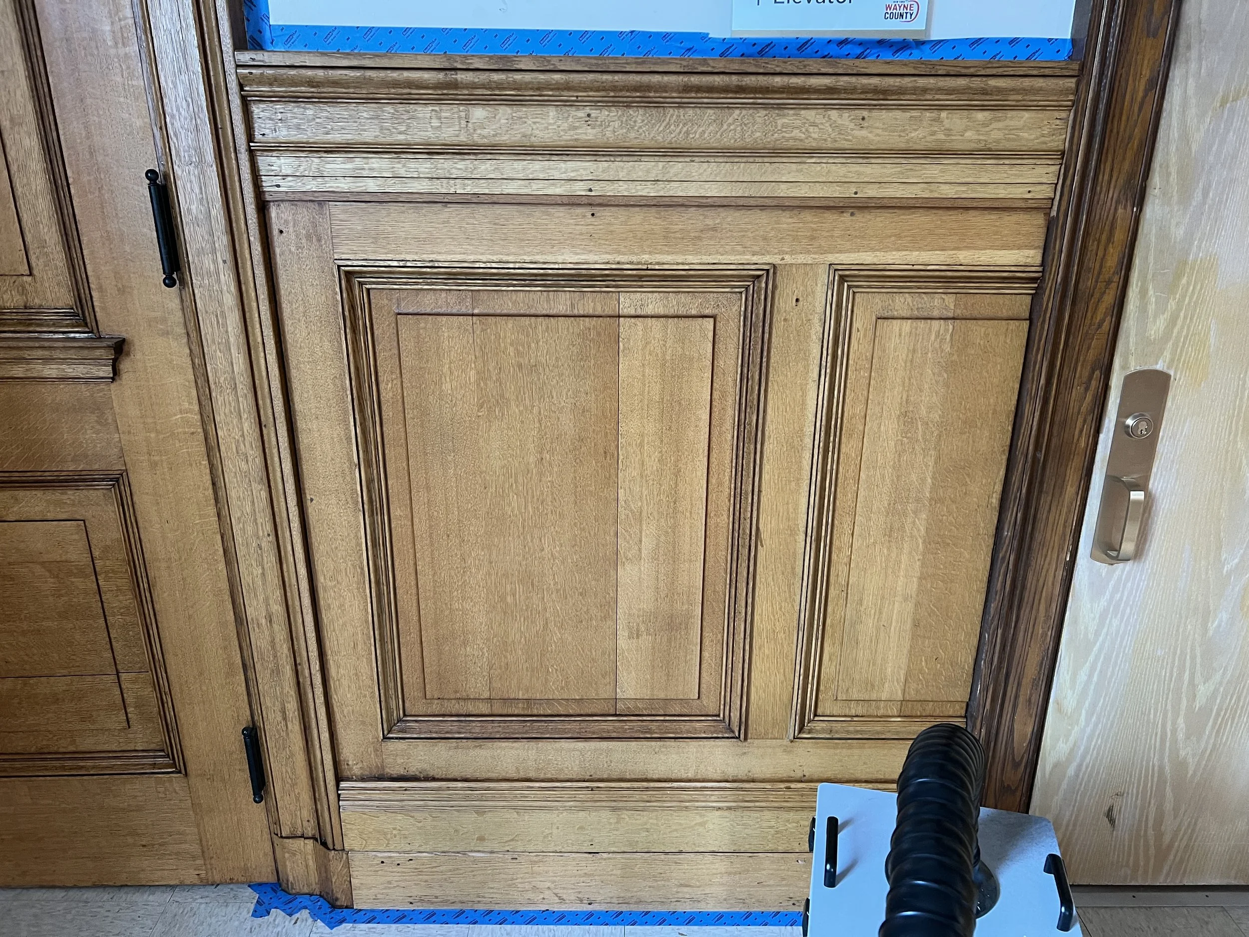 Wooden doors with black handles, in an indoor setting. Part of a person and a piece of equipment are visible at the bottom right corner.