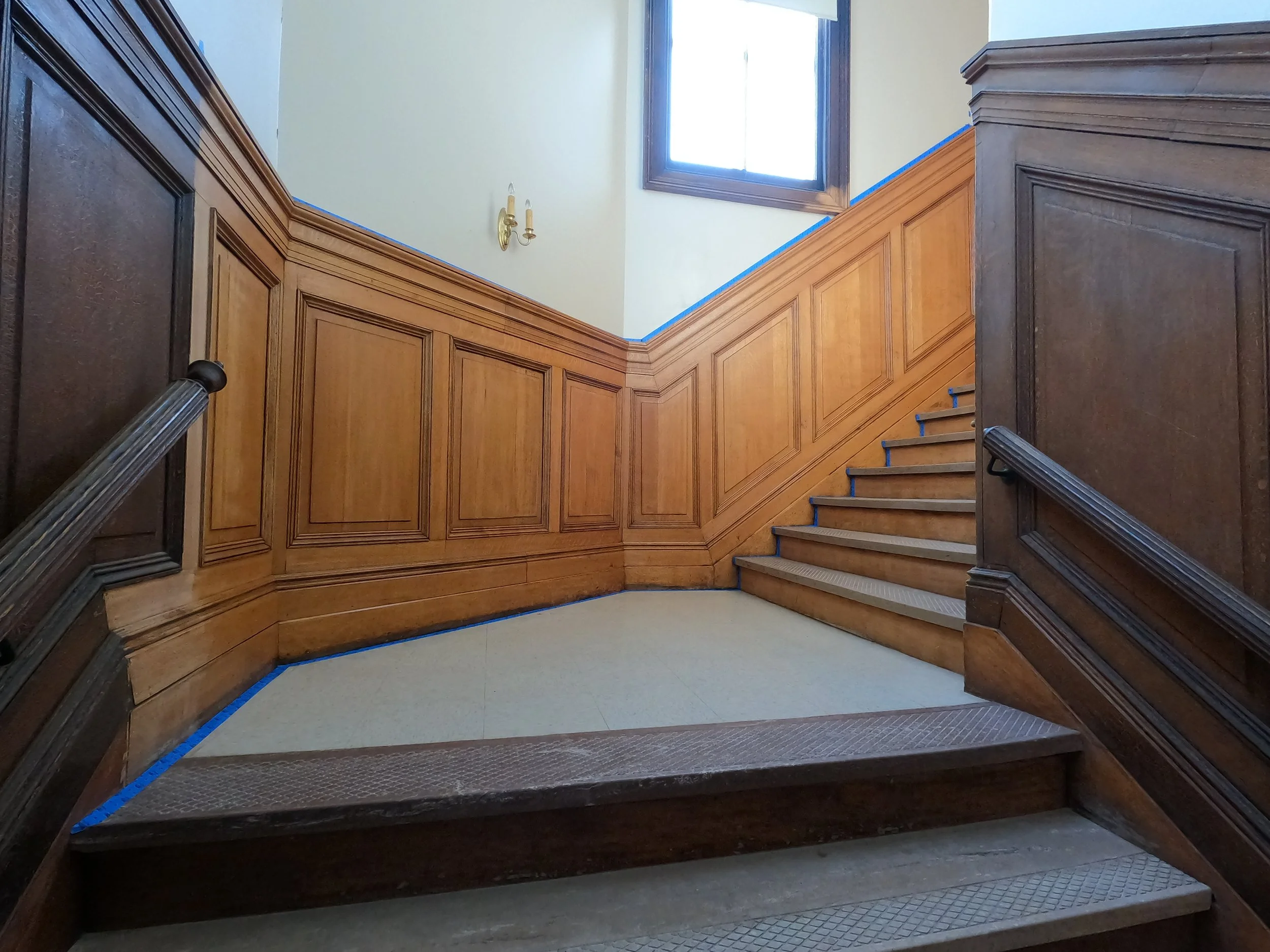 Wood-paneled staircase with wooden walls, a window at the top letting in natural light, and a brass wall sconce, with protective blue painter's tape along the edges of the stairs.