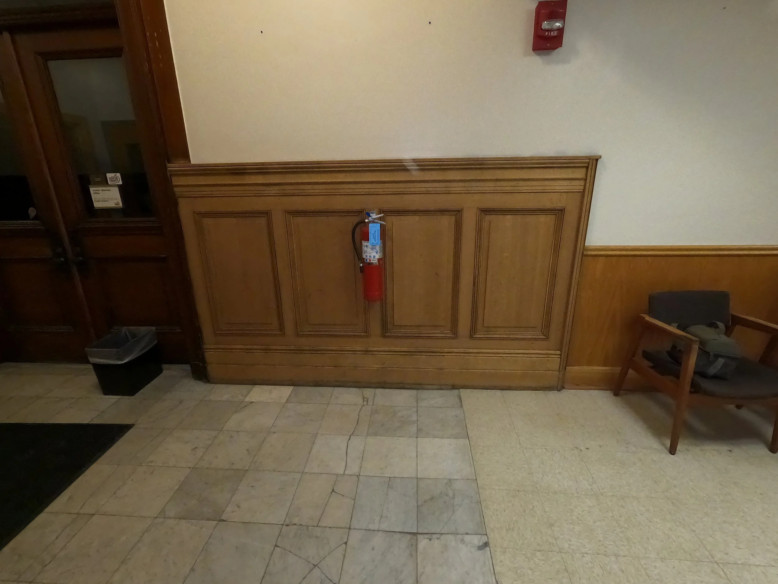 The image shows a beige tiled floor, wooden paneling on the wall, with a fire extinguisher mounted on the wall and a red fire alarm box nearby. There is a black container on the floor and a wooden chair with a black bag on it to the right.