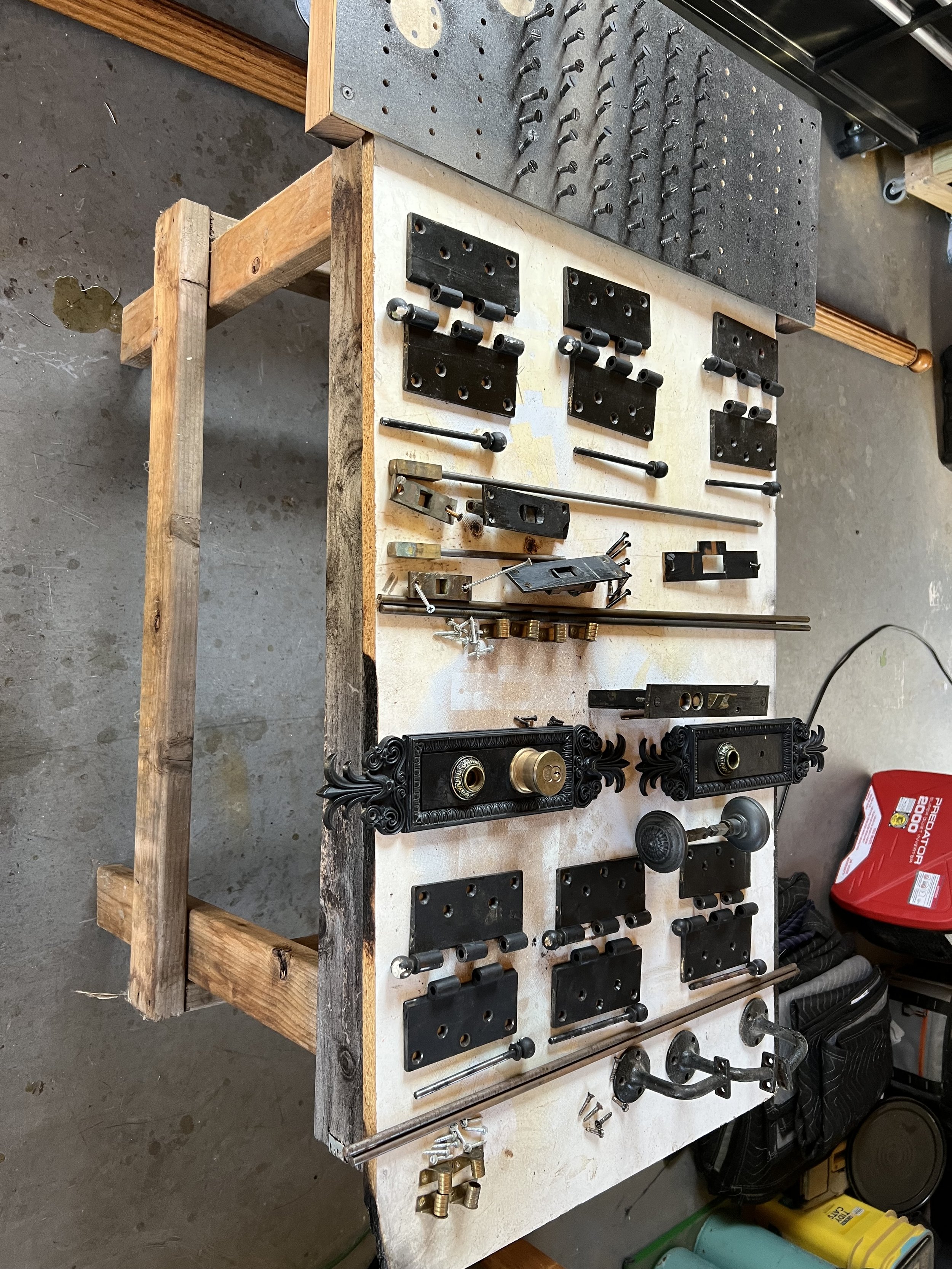A workbench with various disassembled black metallic and brass hardware parts, screws, and tools, with a pegboard above containing additional hardware, in a workshop or garage setting.