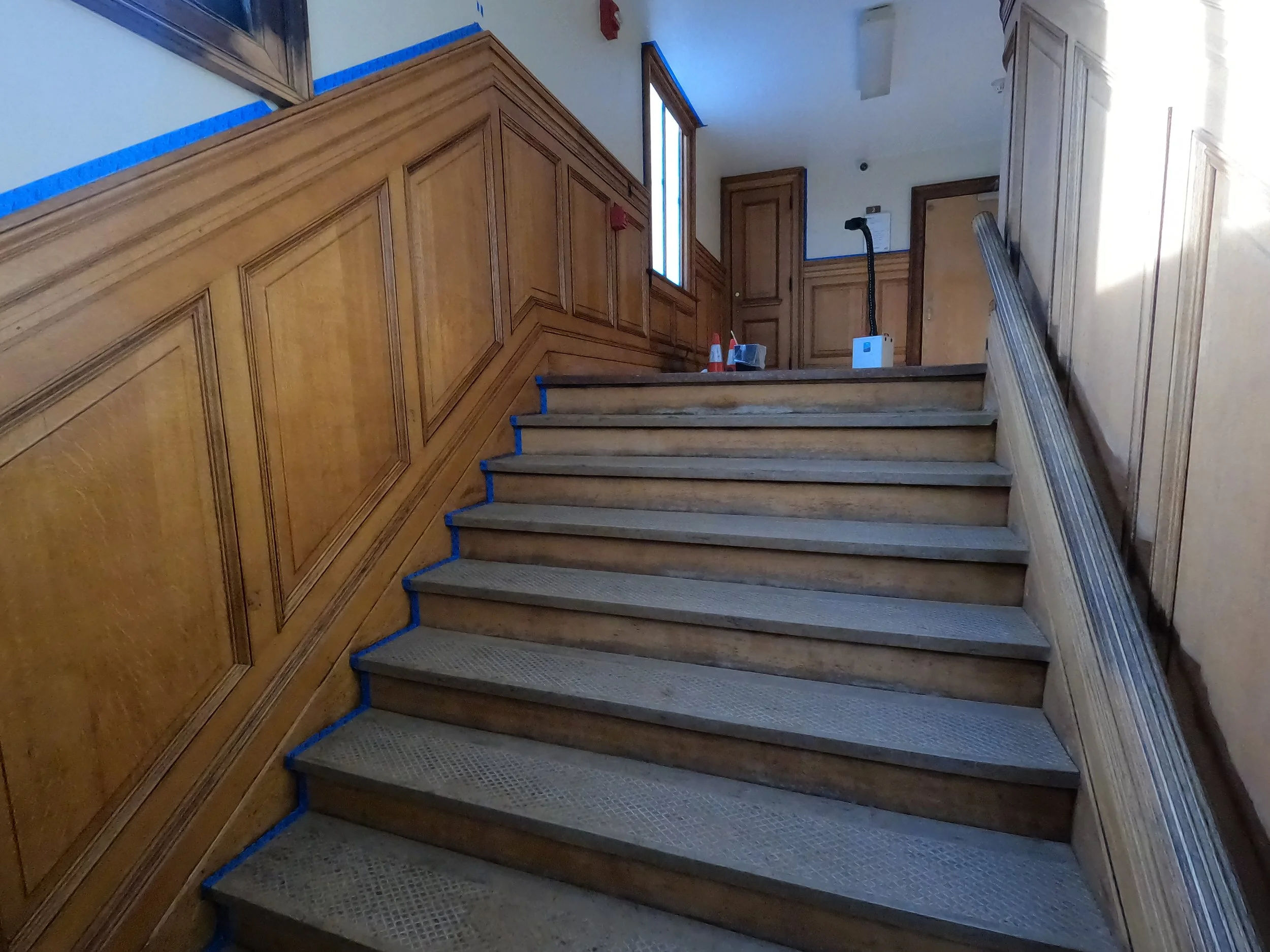 Wooden staircase with blue tape along the edges of each step and wooden wall paneling on the side. Construction tools and cones are placed at the top.