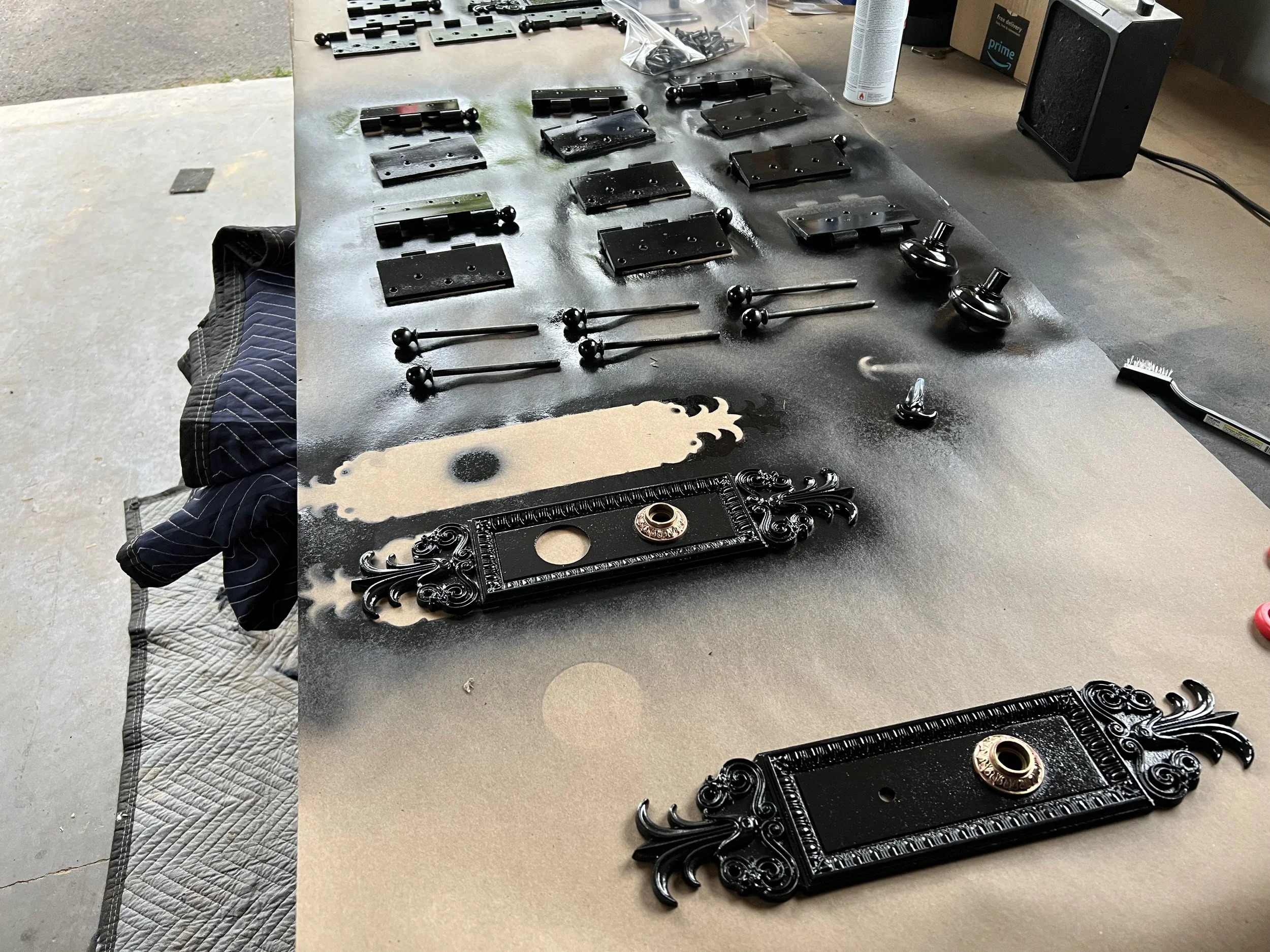 A workbench with black decorative cast iron frames, some with ornate designs, and metal hardware. Items include metal hinges, a screwdriver, and other small parts, with a spray can, a black speaker, and cardboard boxes in the background.