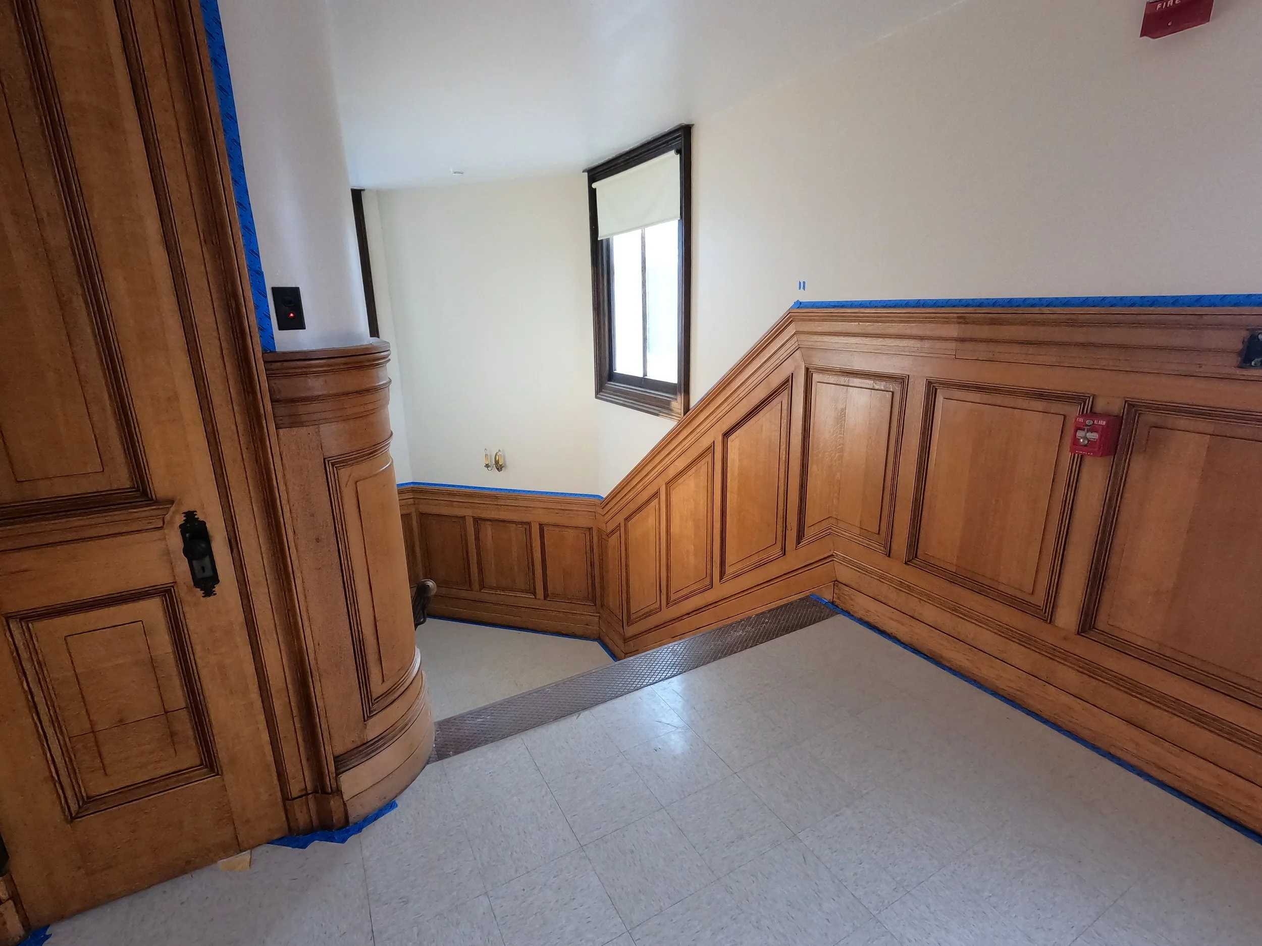 Interior of a stairwell with wooden paneling on walls and crane framing a window, with blue painter's tape along edges and a fire alarm on the wall.