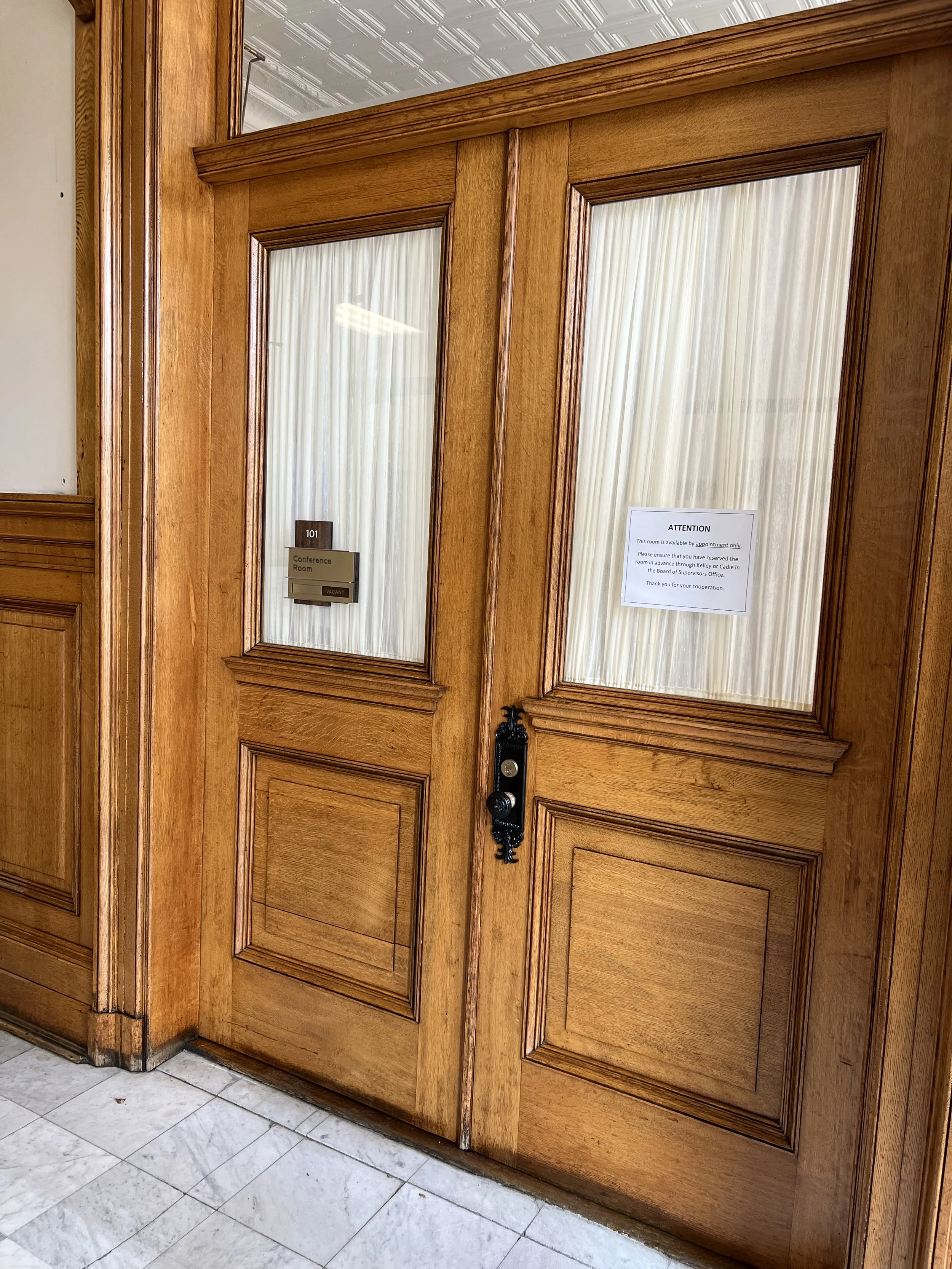 Wooden double doors with glass panels covered by white curtains, a sign on the right door indicates the room is available by appointment only, located inside a building with a tiled floor.