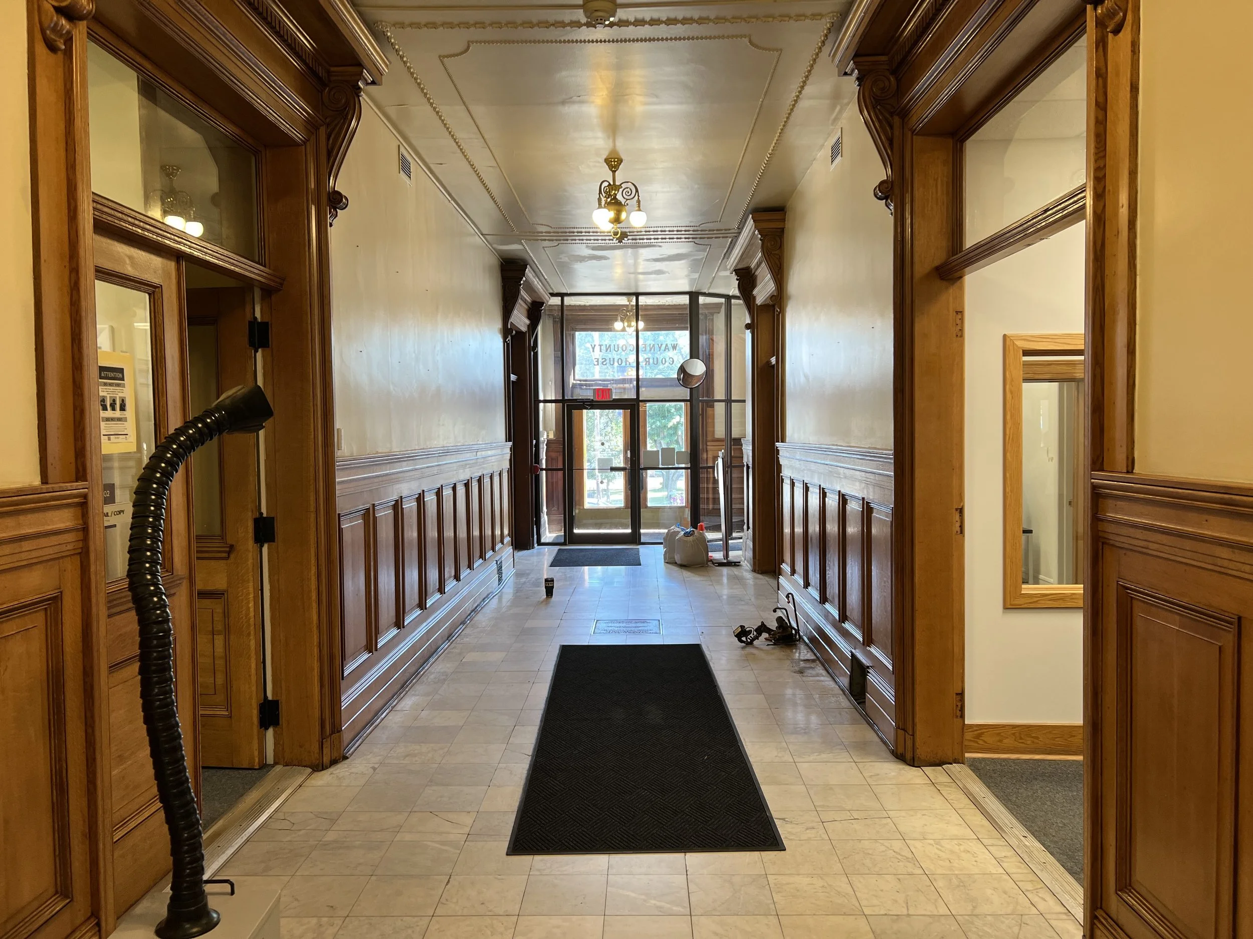 A hotel lobby with a tiled floor and wood-paneled walls on either side, leading to a glass door entrance. There are ceiling lights and some construction or cleaning supplies on the floor near the entrance.