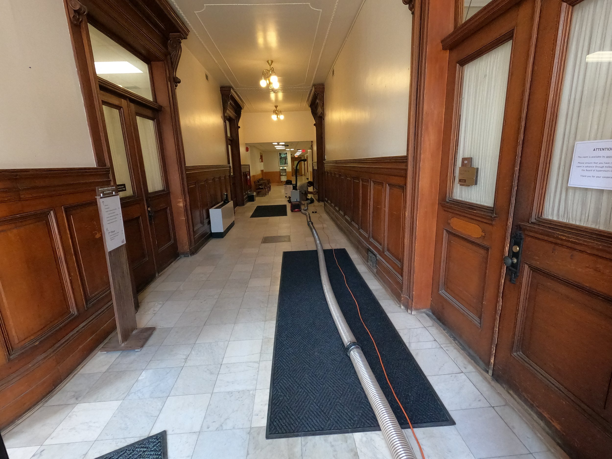 Hallway with wood-paneled walls and tiled floor, pipe or duct on the floor, ceiling lights, and signs on the walls.