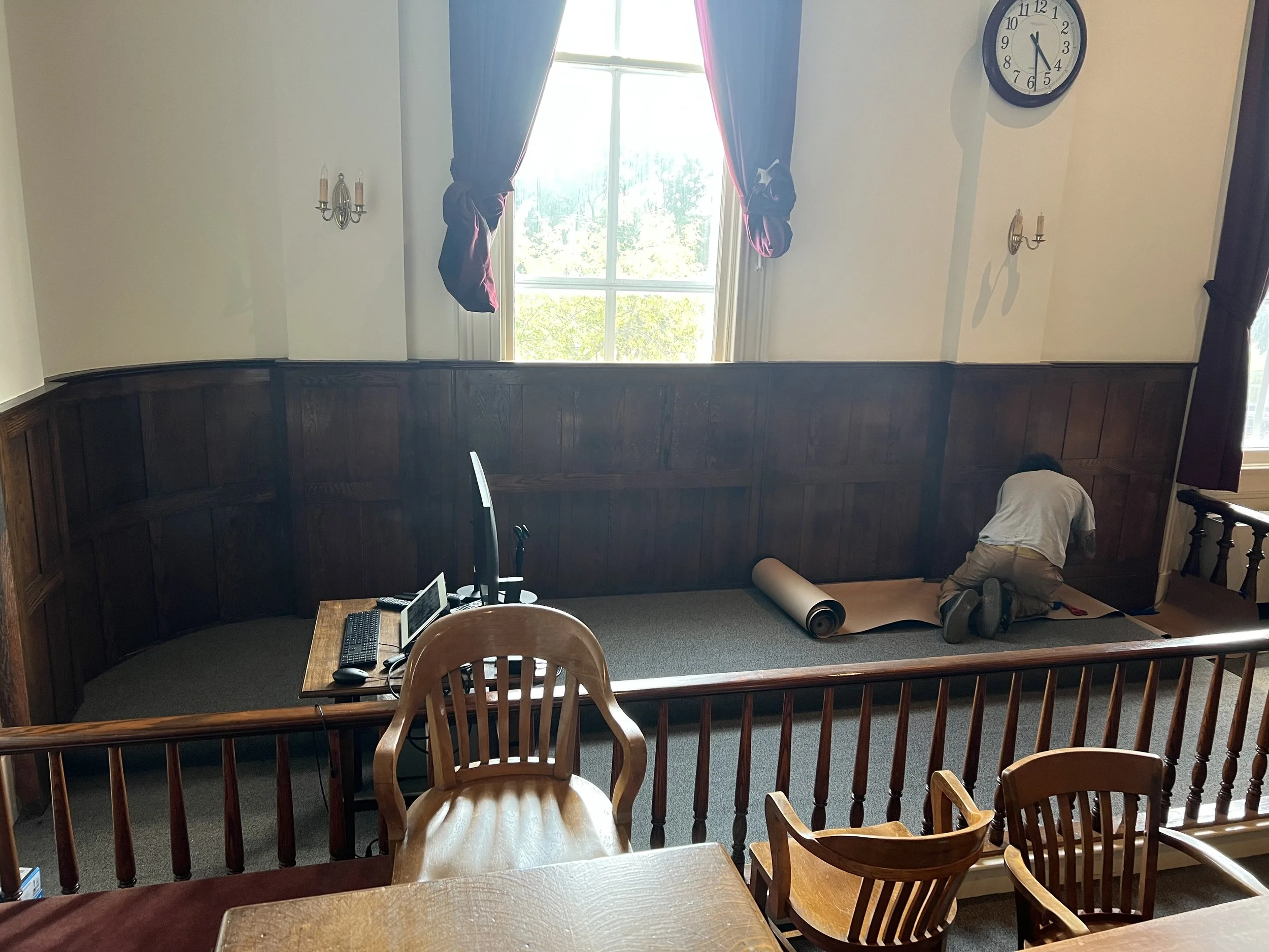 A person kneeling on the floor assembling or repairing something in a room with a large window, wood-paneled lower wall, and two wall sconces. There is a wooden table with a monitor, keyboard, and mouse in the foreground, and a wooden chair with a curved backrest nearby.