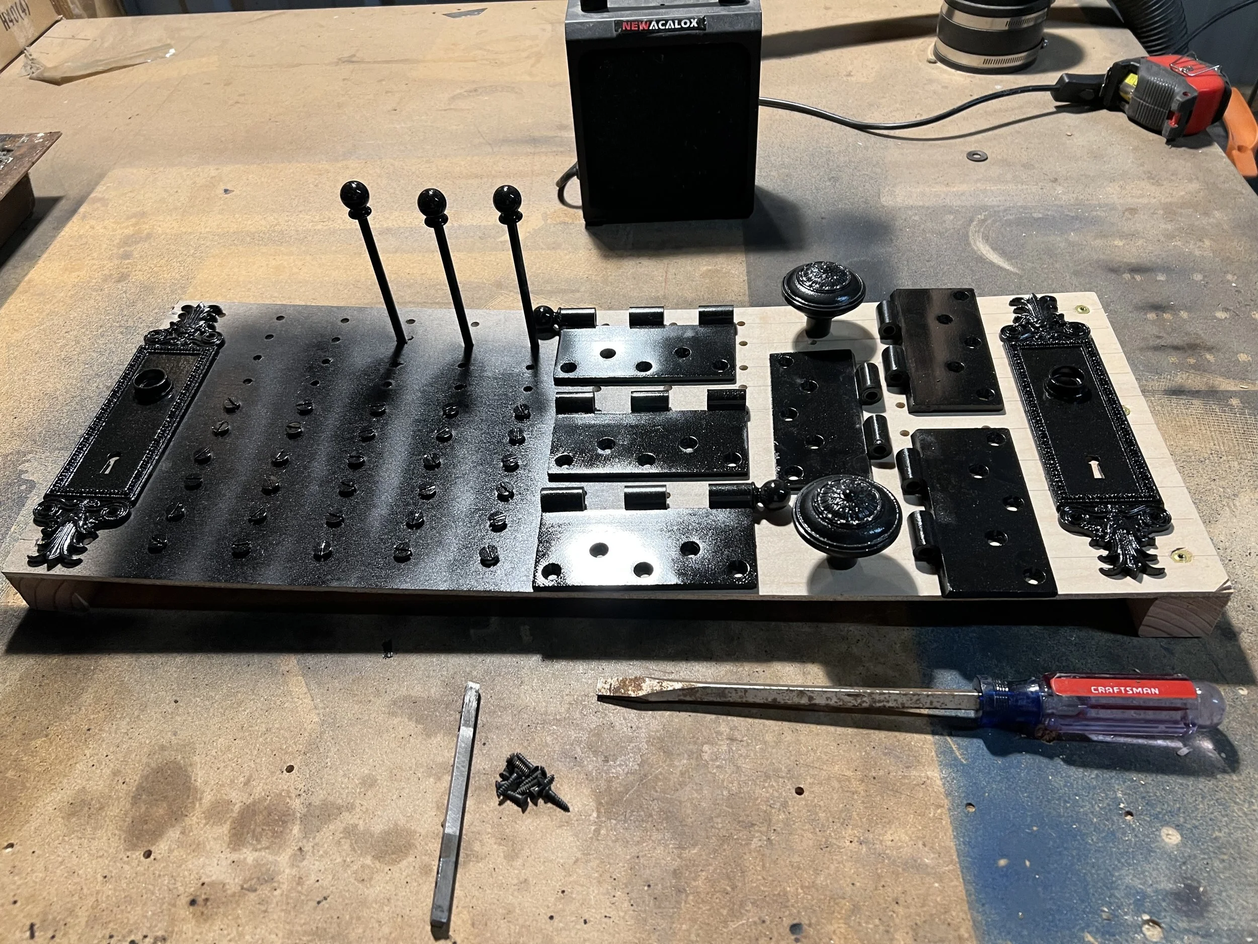 A woodworking workbench with black metal decorative items, various tools, and parts arranged on a white wooden board. A cordless screwdriver and a measuring tape are visible, and the background shows a dusty workshop setting.