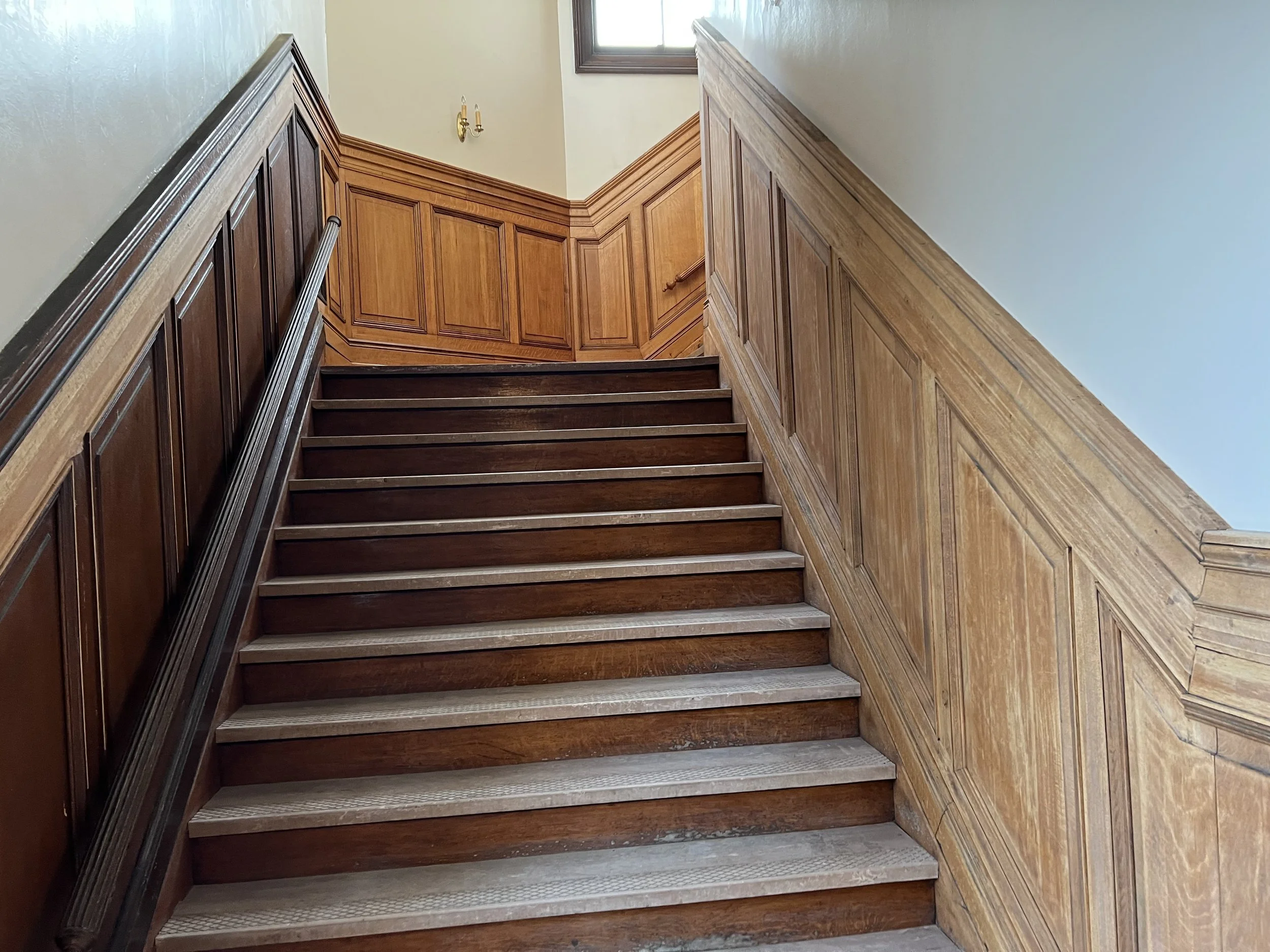 Wooden staircase with handmade wainscoting on the walls, a brass wall sconce with two candles, and a small window at the top of the staircase letting in natural light.