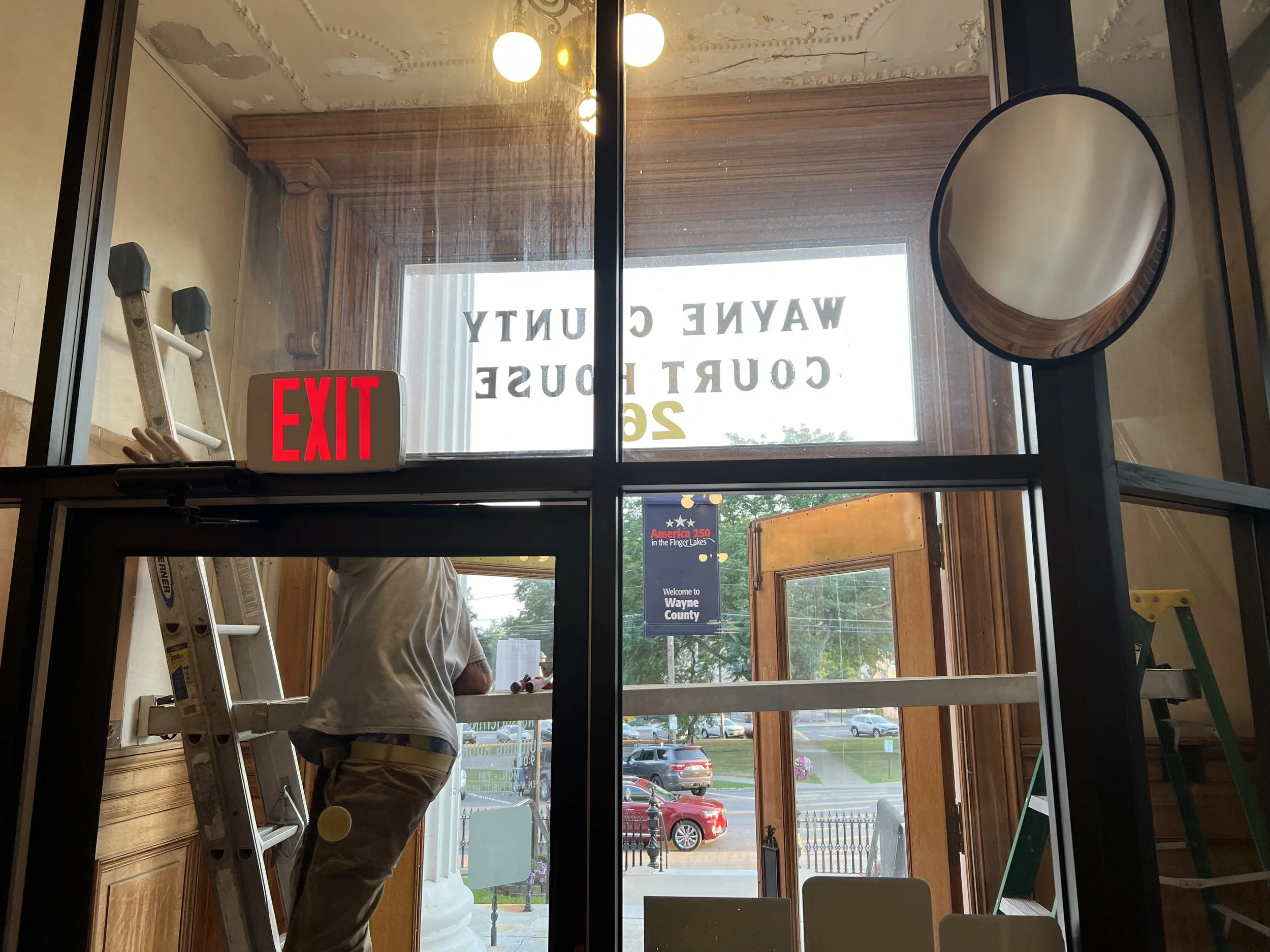 Inside a building, a person is on a ladder cleaning or repairing a wooden doorway or window frame near the glass door. An illuminated red 'EXIT' sign is visible above a glass entrance door. The glass door has text that reads 'WAYNE COUNTY HUNTY' and ‘22’. Seen through the glass door are parked cars and a parking lot outside. The interior has wooden finishing and a round mirror on the wall.