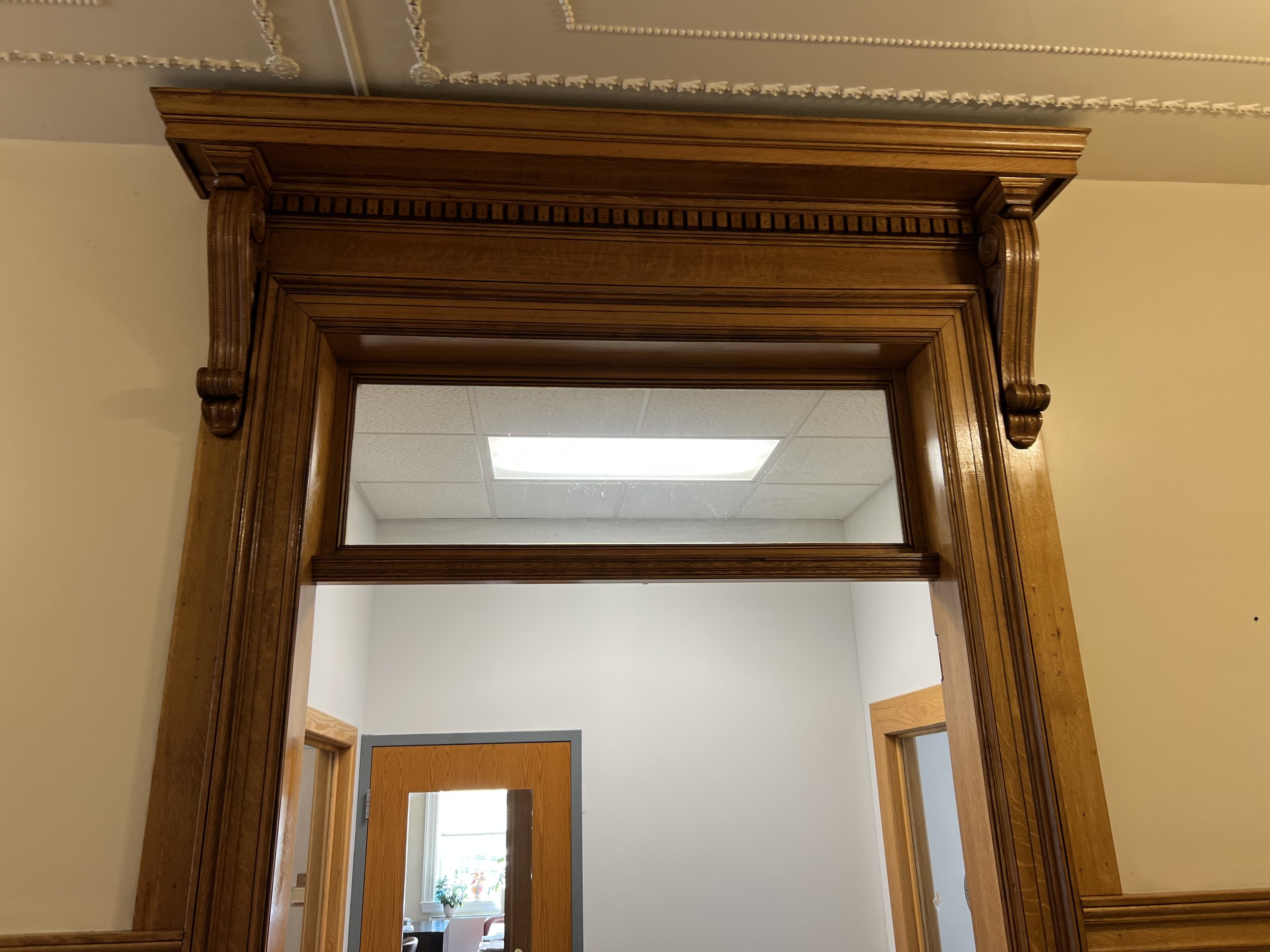 Interior of a room showing a large, ornate wooden doorway with decorative molding. Through the doorway, a window with a small potted plant on the windowsill is visible, letting in natural light.