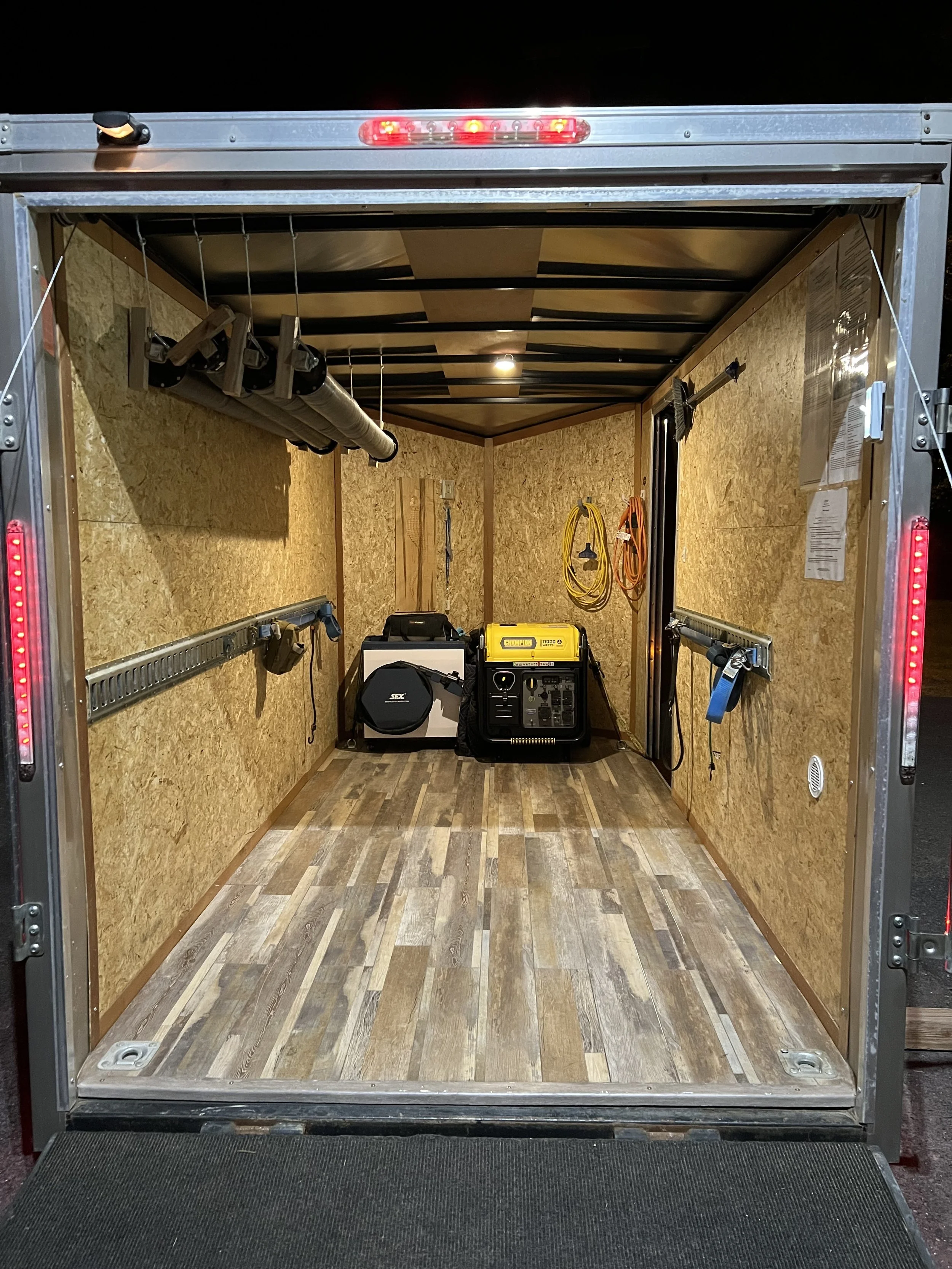 Empty industrial trailer with wooden flooring and walls, tools, extension cords, and equipment inside, night setting.